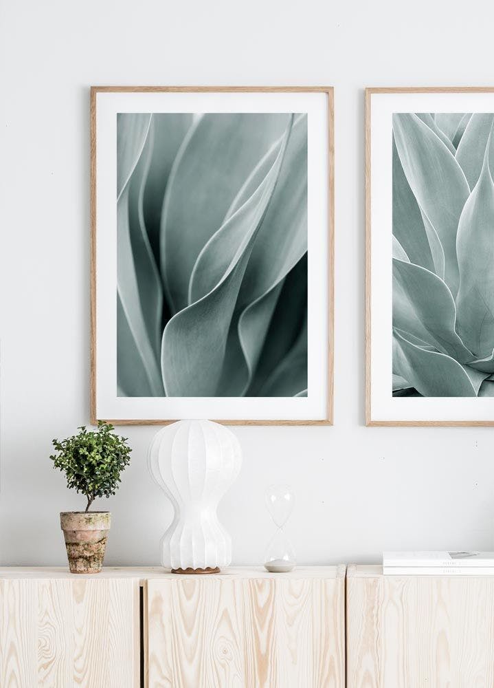 Two posters of abstract green agave plant leaves hang above a light wood cabinet with a small plant, white lamp, and hourglass.