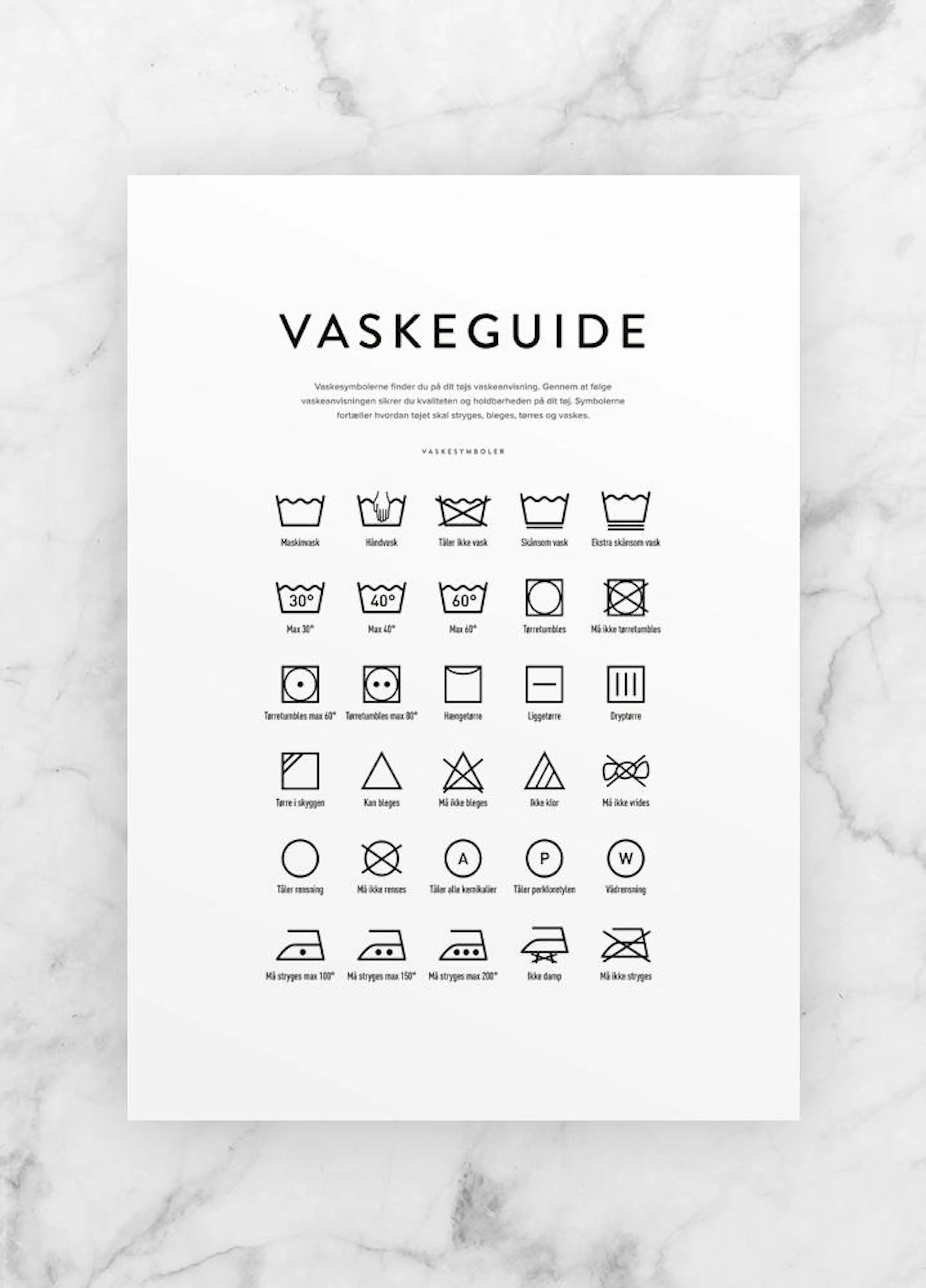 Plakát s průvodcem symbolů praní prádla v bílé a černé barvě, položený na bílém mramorovém povrchu.