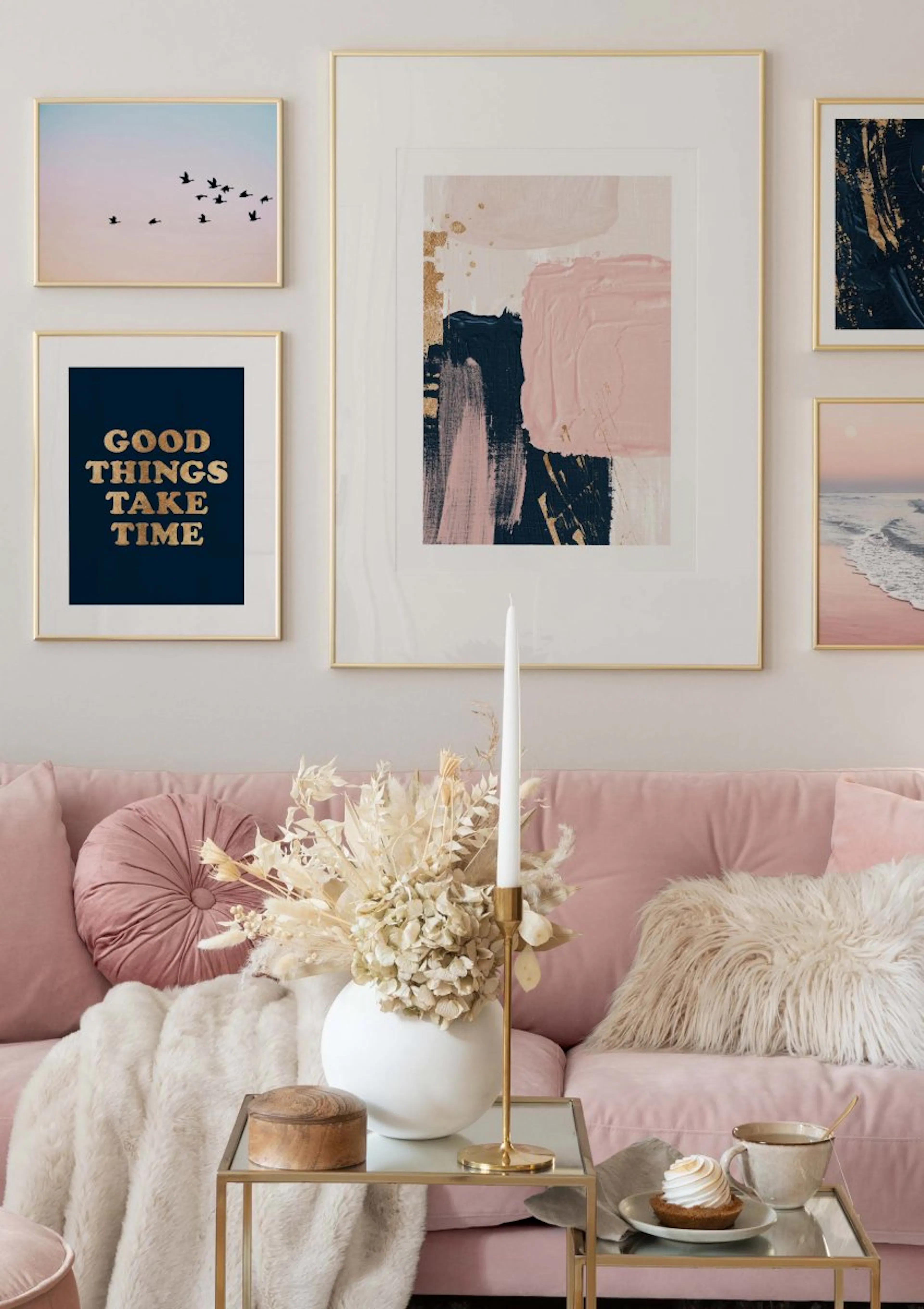 ピンクのソファの上に飾られたギャラリーウォール。抽象的なポスター、鳥の群れ、青い波の風景、そして「GOOD THINGS TAKE TIME」という文字のポスターが見える。
