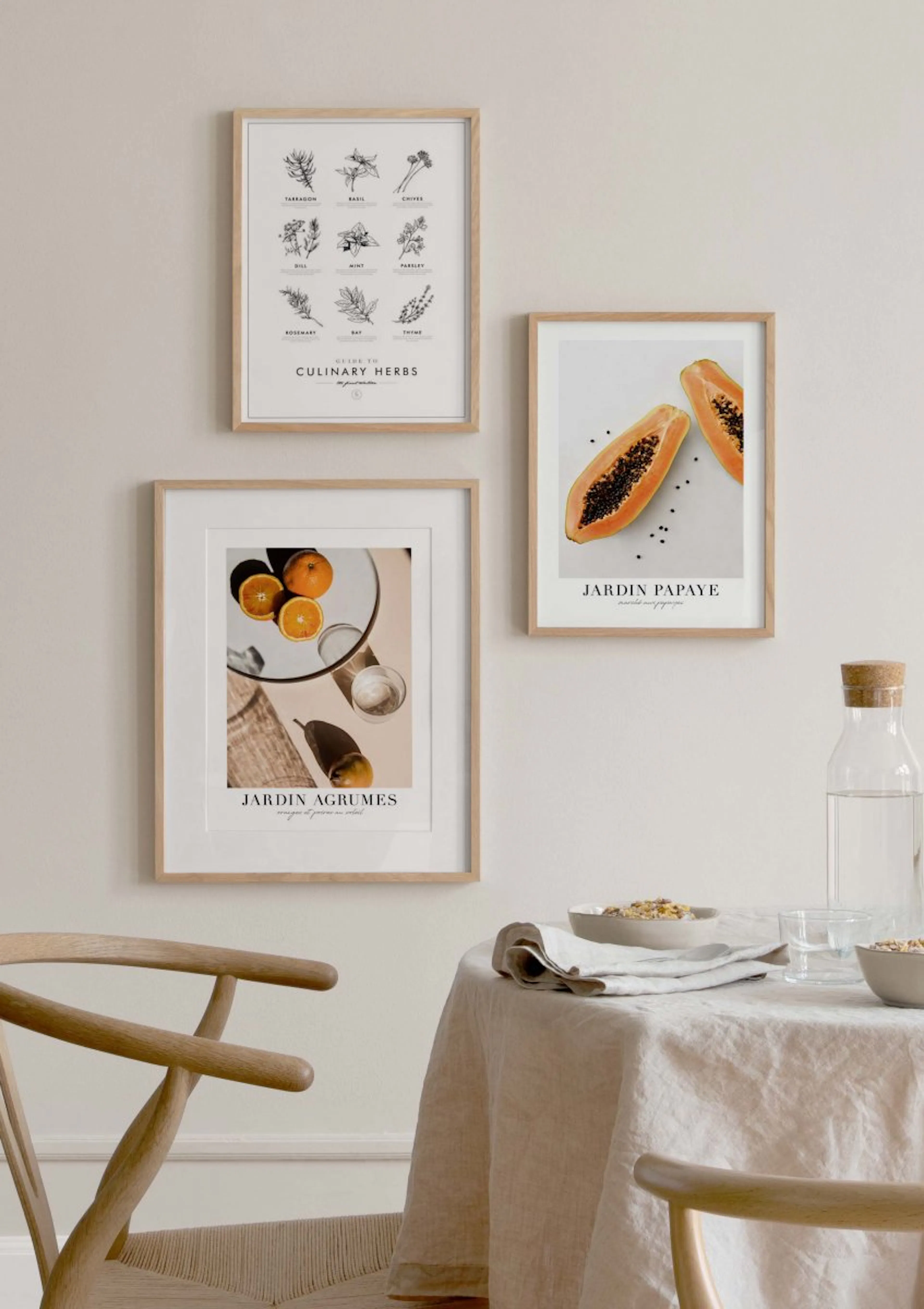 A gallery wall of three botanical and fruit posters above a dining table with a linen tablecloth and water bottle.
