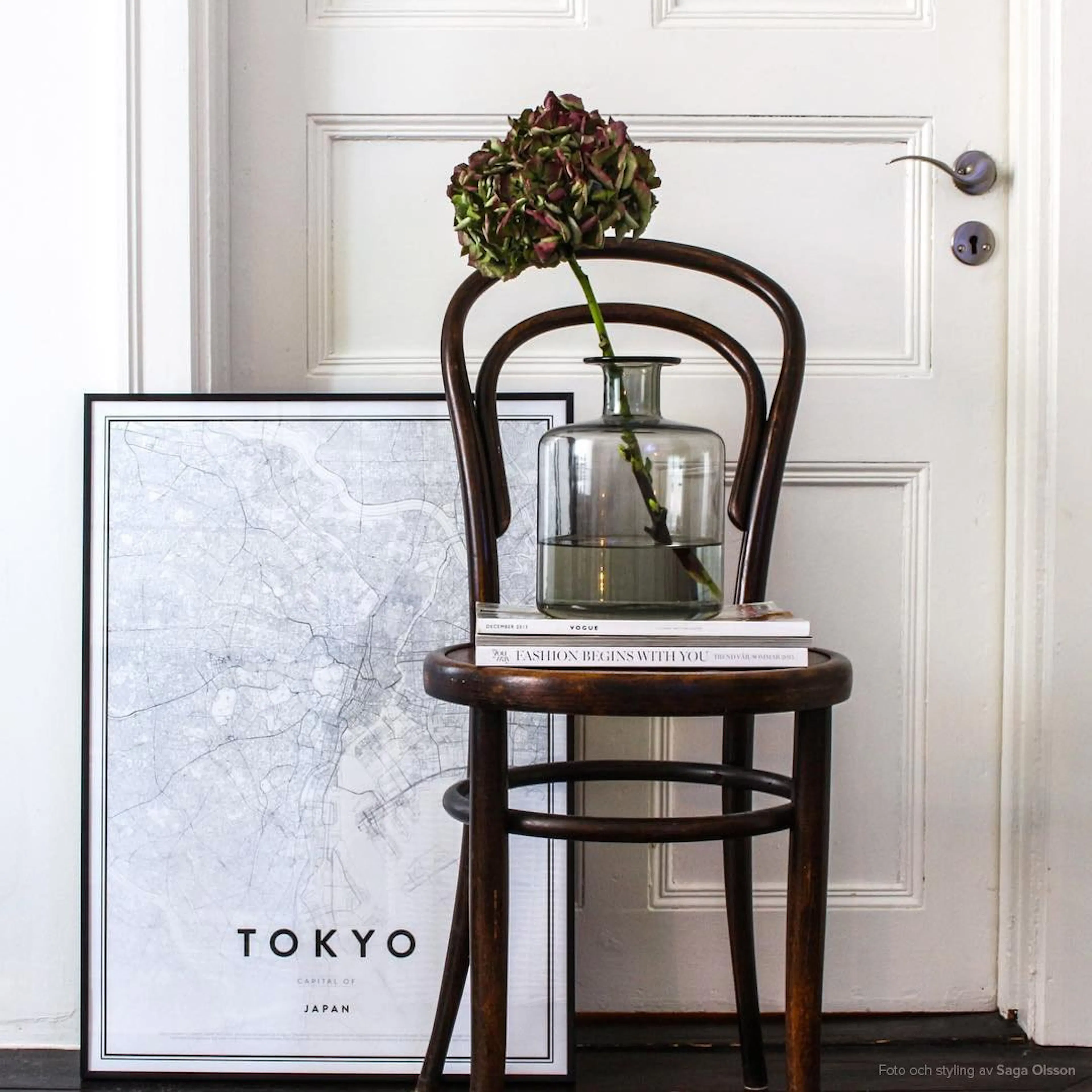 A black and white Tokyo map poster leaning against a white door, beside a wooden chair with a vase and books.