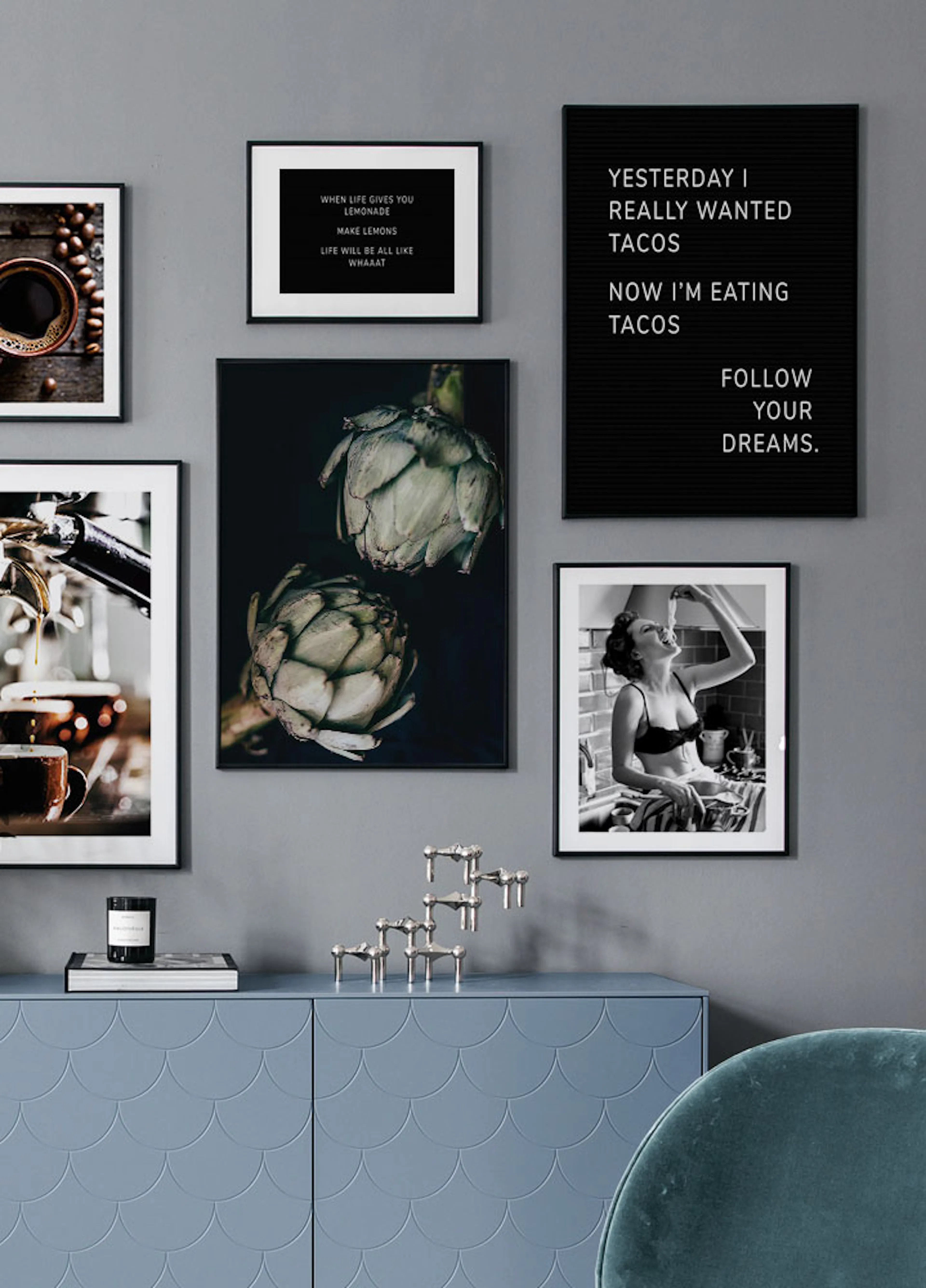 Julisteseinä harmaalla seinällä, sinisen lipaston yläpuolella. Julisteissa on kahvikuppi, lattekone, kaksi artisokkaa, tekstiä j