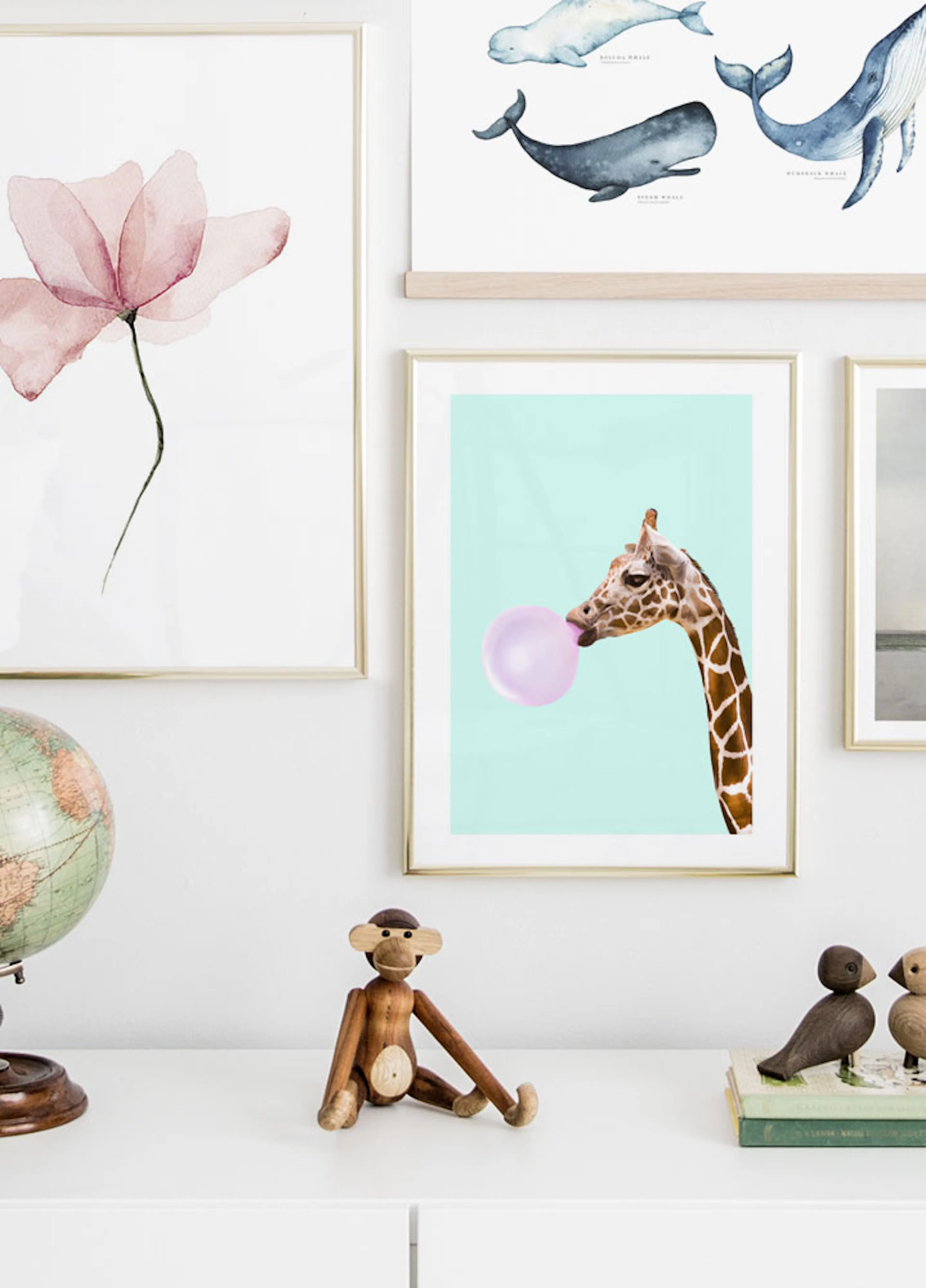 A giraffe with a pink bubblegum bubble on a mint background, part of a gallery wall above white furniture.