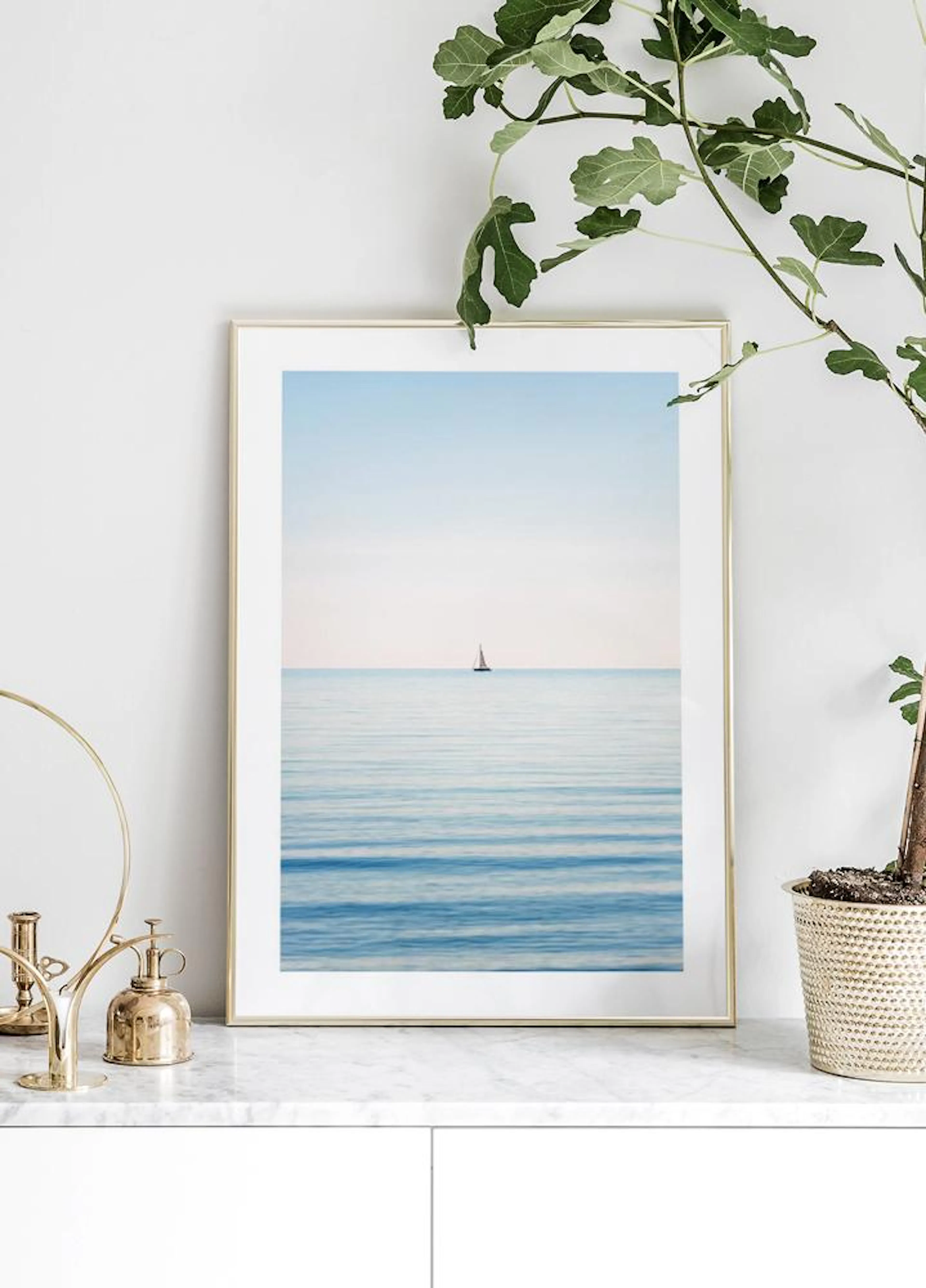 A nautical poster of a lone sailboat on calm blue ocean waters, displayed on a marble countertop with a plant and brass decor.