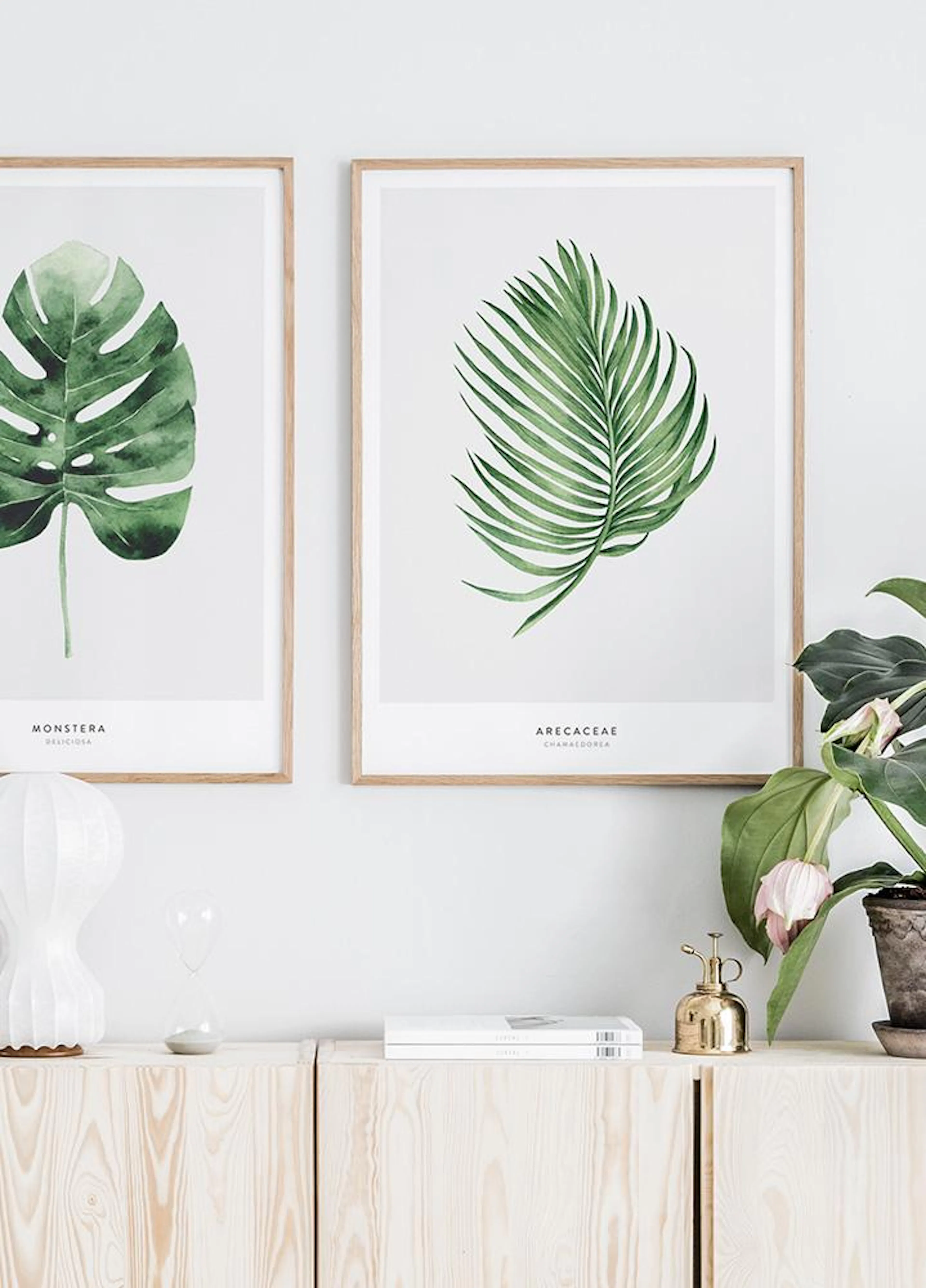 Due poster con illustrazioni di foglie verdi, Monstera e Areca, appesi sopra una credenza in legno chiaro con decorazioni.