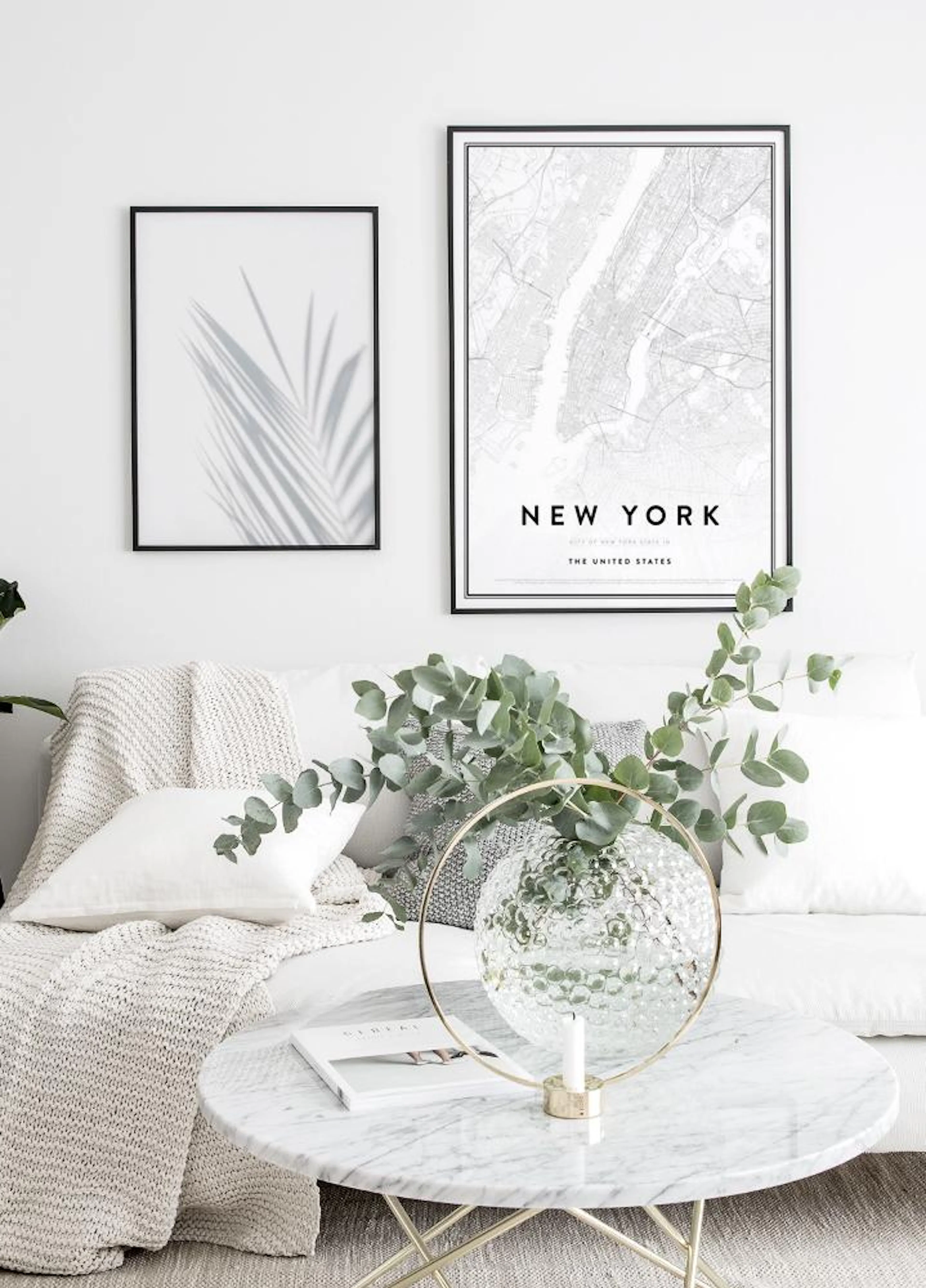 A black and white New York city map poster and a botanical palm leaf shadow print hanging above a white sofa in a modern living 