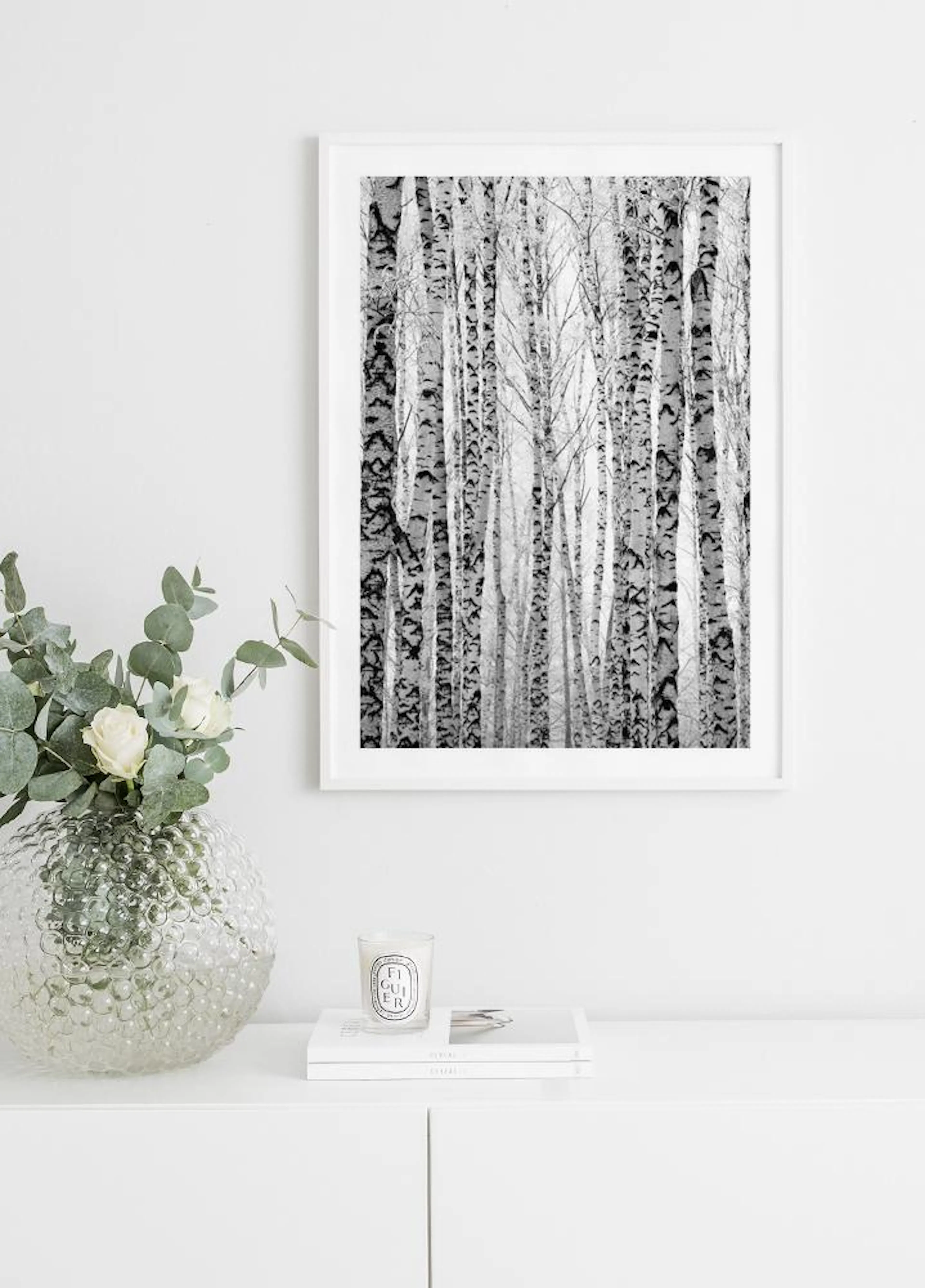A black and white birch forest poster displayed above a white cabinet with a vase of flowers and a candle.