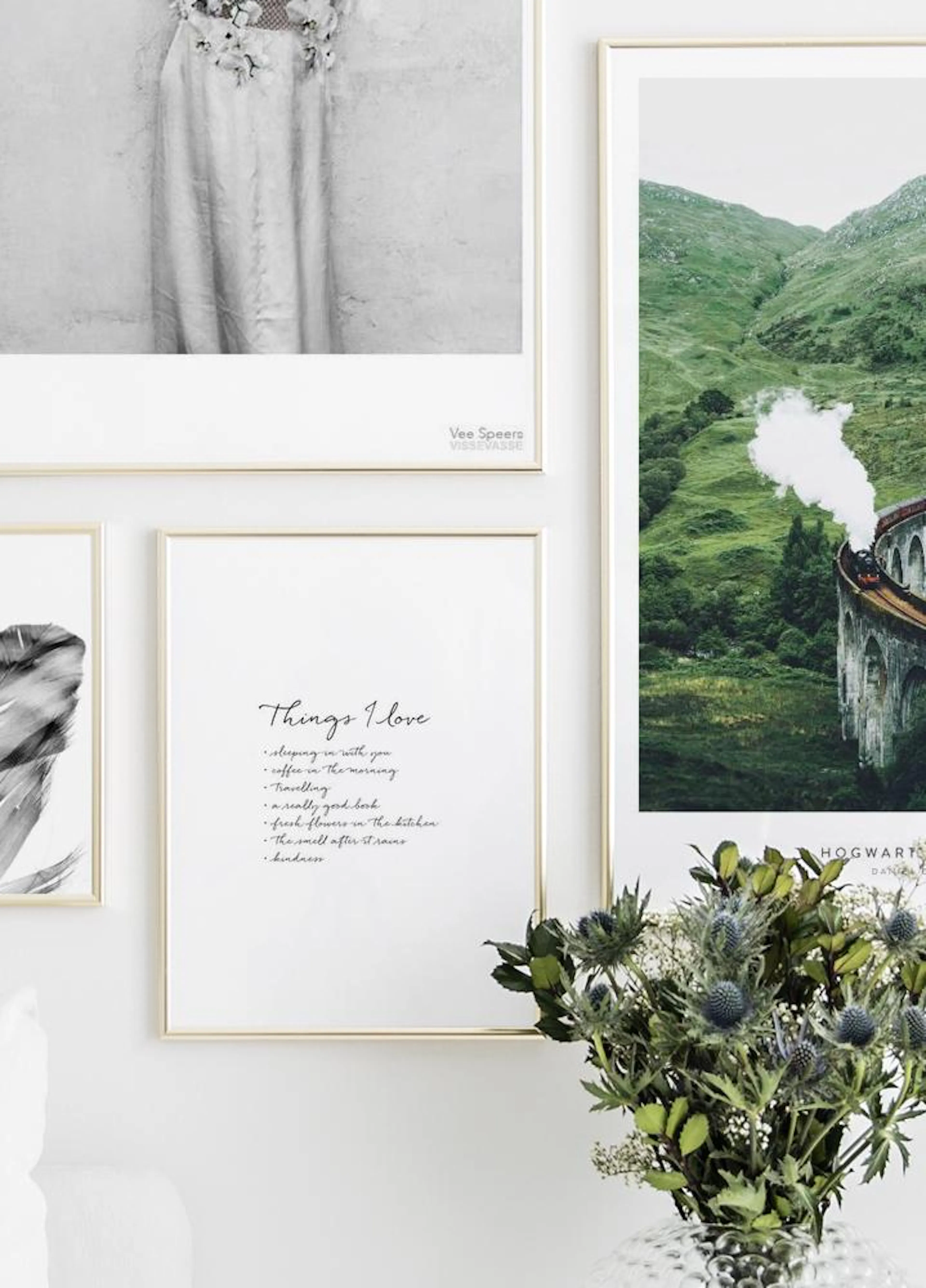白い壁に飾られたポスター。蒸気を上げて走る列車が緑の山並みの中の古い橋を渡るポスター、羽のポスター、そして手書き風の書体で「Things I Love」と書かれたポスターが並ぶ。手前には花瓶に生けられた青いアザミと緑の葉が見える。