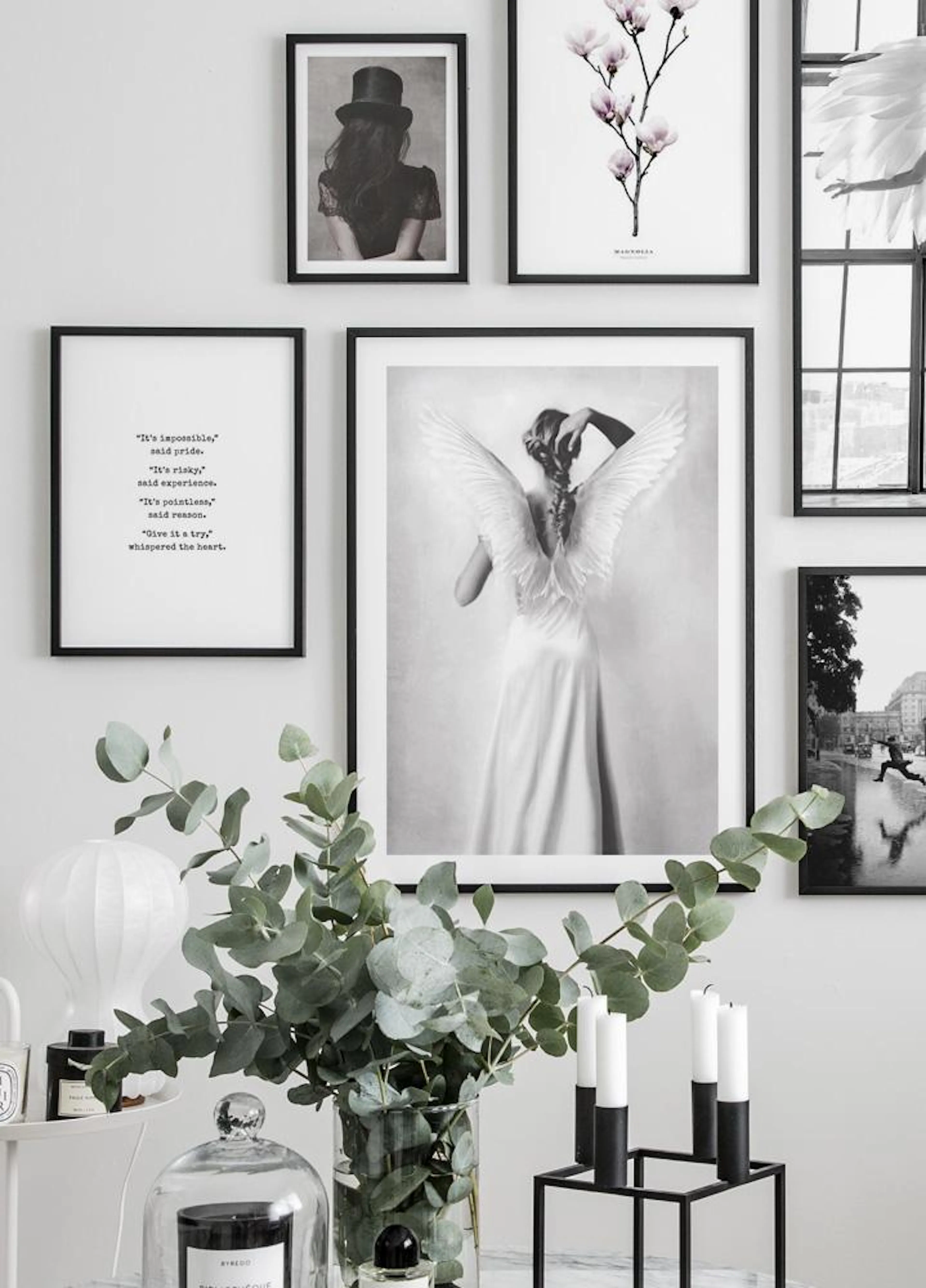 A gallery wall with black and white and colour prints, including a woman with wings, a magnolia, and text, displayed above a vas