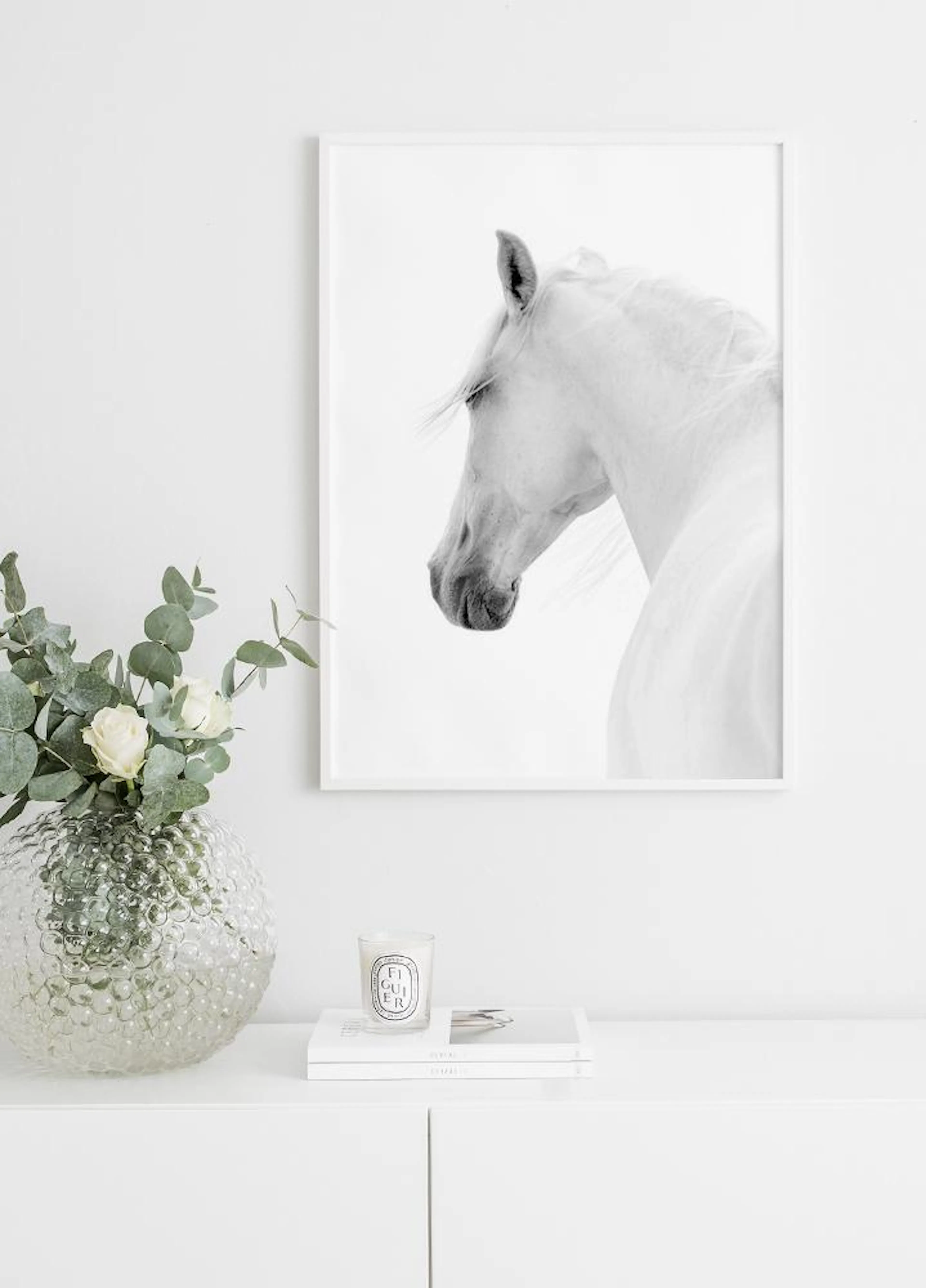 Mustavalkoinen juliste valkoisesta hevosesta, joka on esillä valkoisen lipaston päällä, vieressään maljakko ja kynttilä.