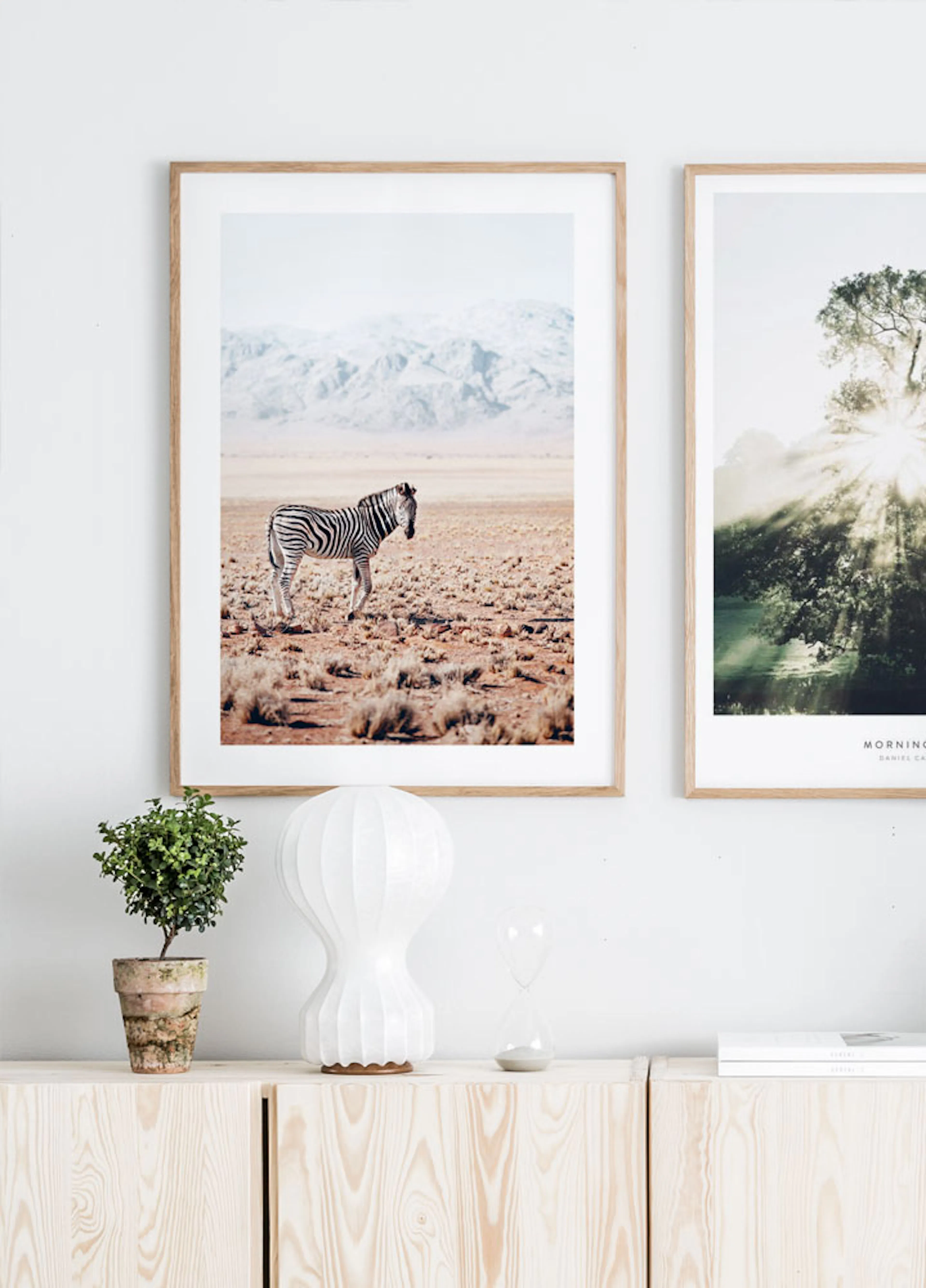 A zebra in a vast, dry landscape with mountains in the background, displayed as a poster above a light wooden cabinet with decor