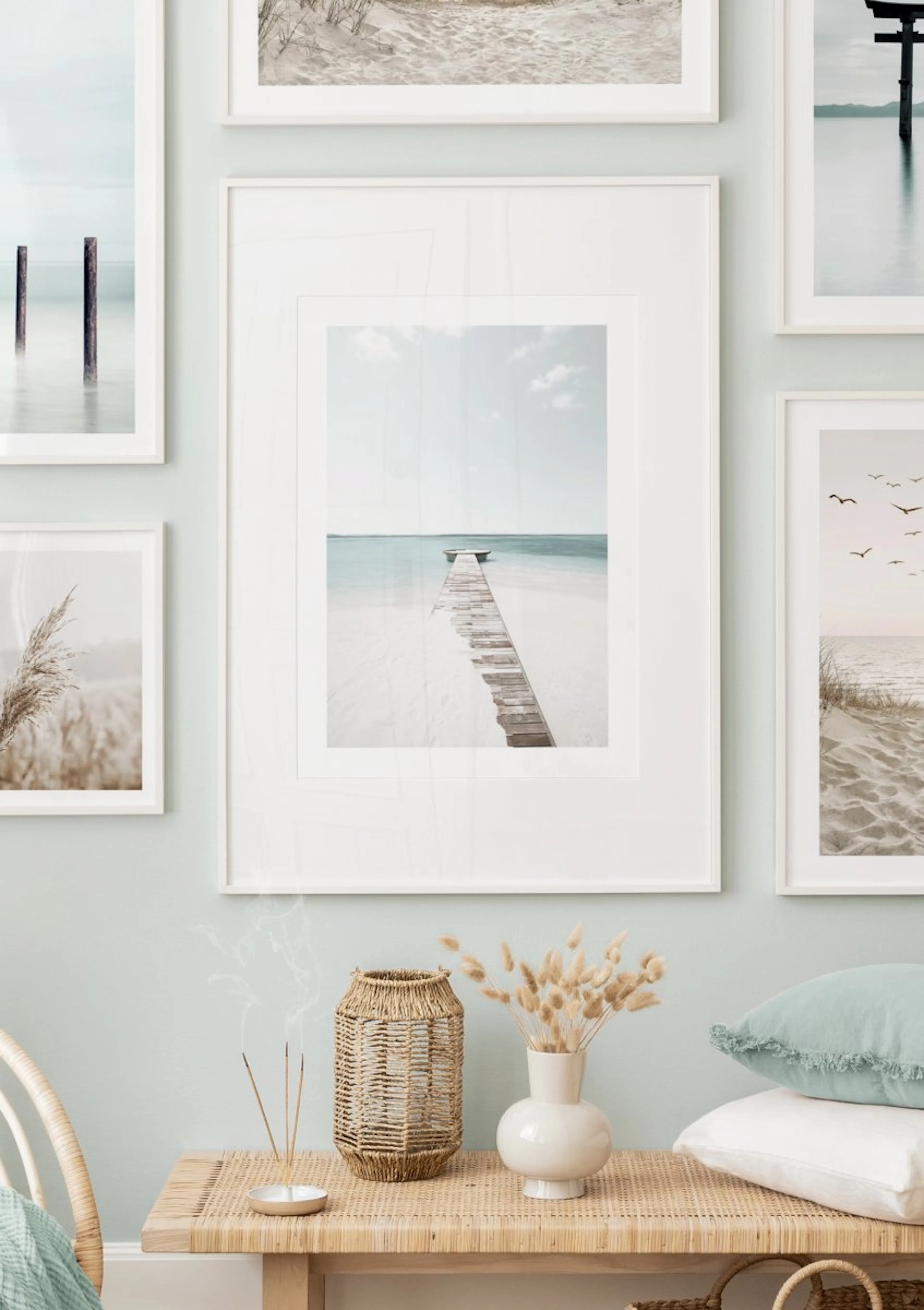 A gallery wall featuring five coastal posters in a light blue room, with a wooden bench, wicker basket, and dried flowers below.