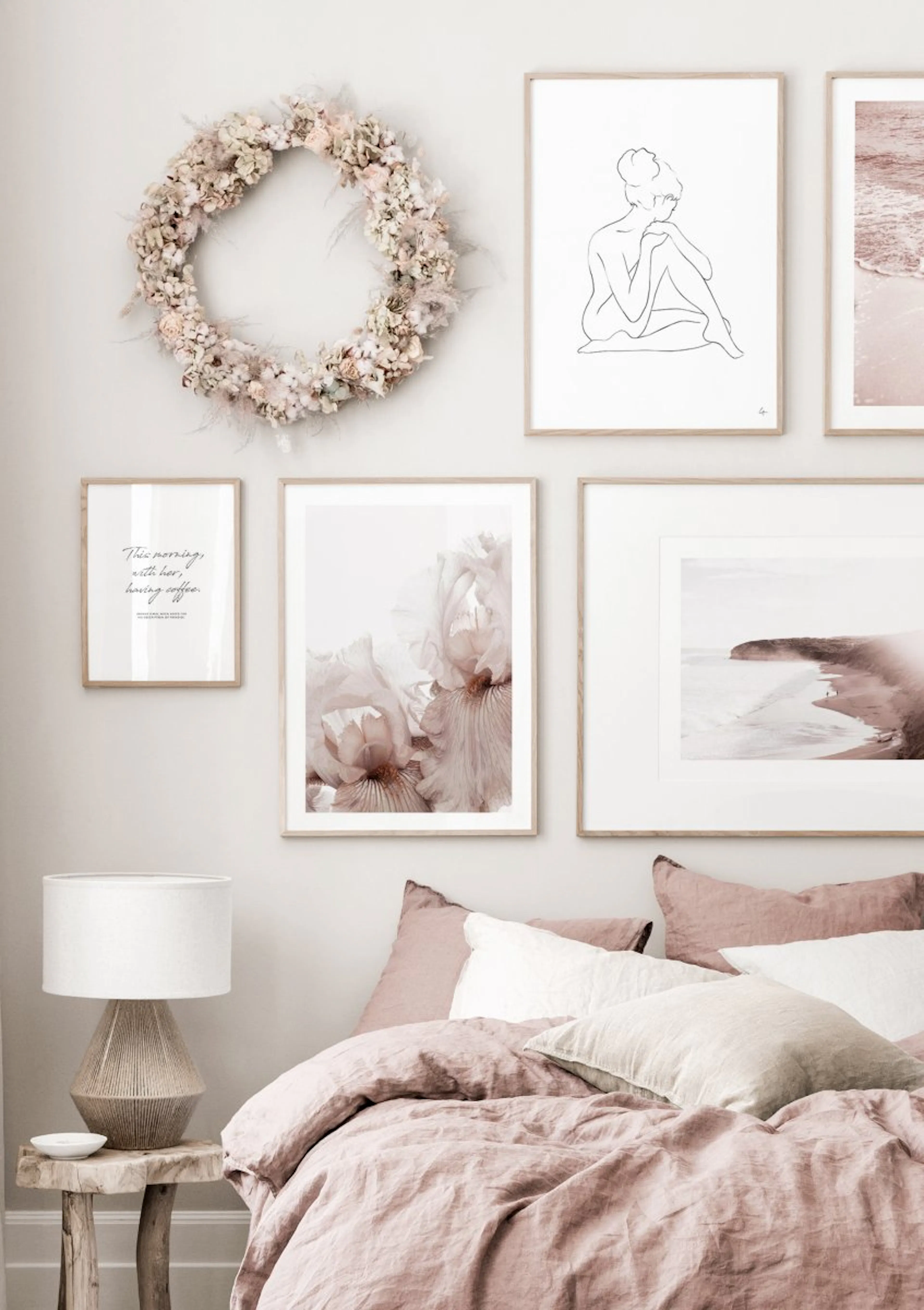 A gallery wall with posters including a line art figure, pale pink irises, and a misty beach, displayed above a bed with pink li