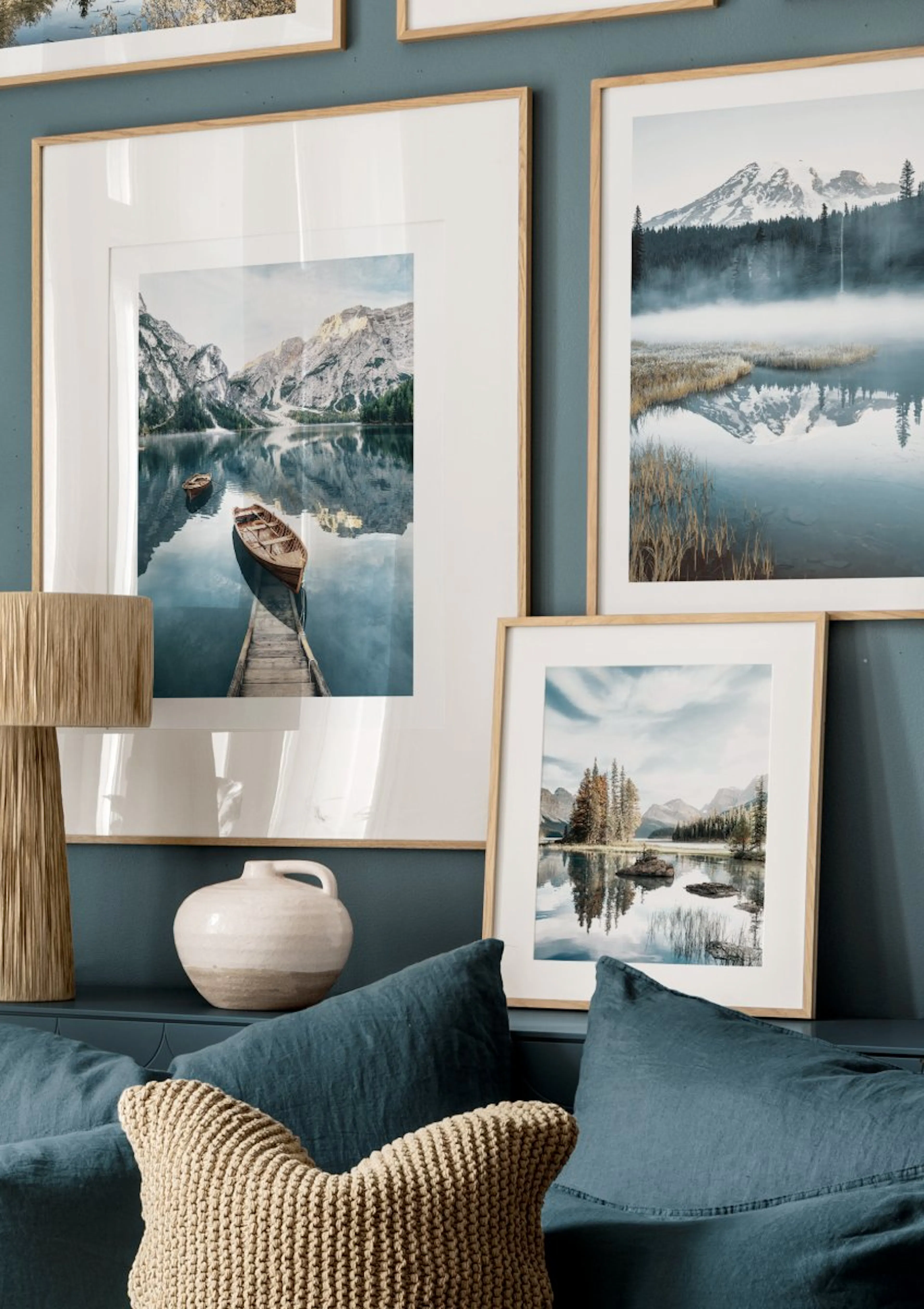 A gallery wall featuring tranquil lake and mountain landscape posters, with blue cushions and a woven lamp in a living room.