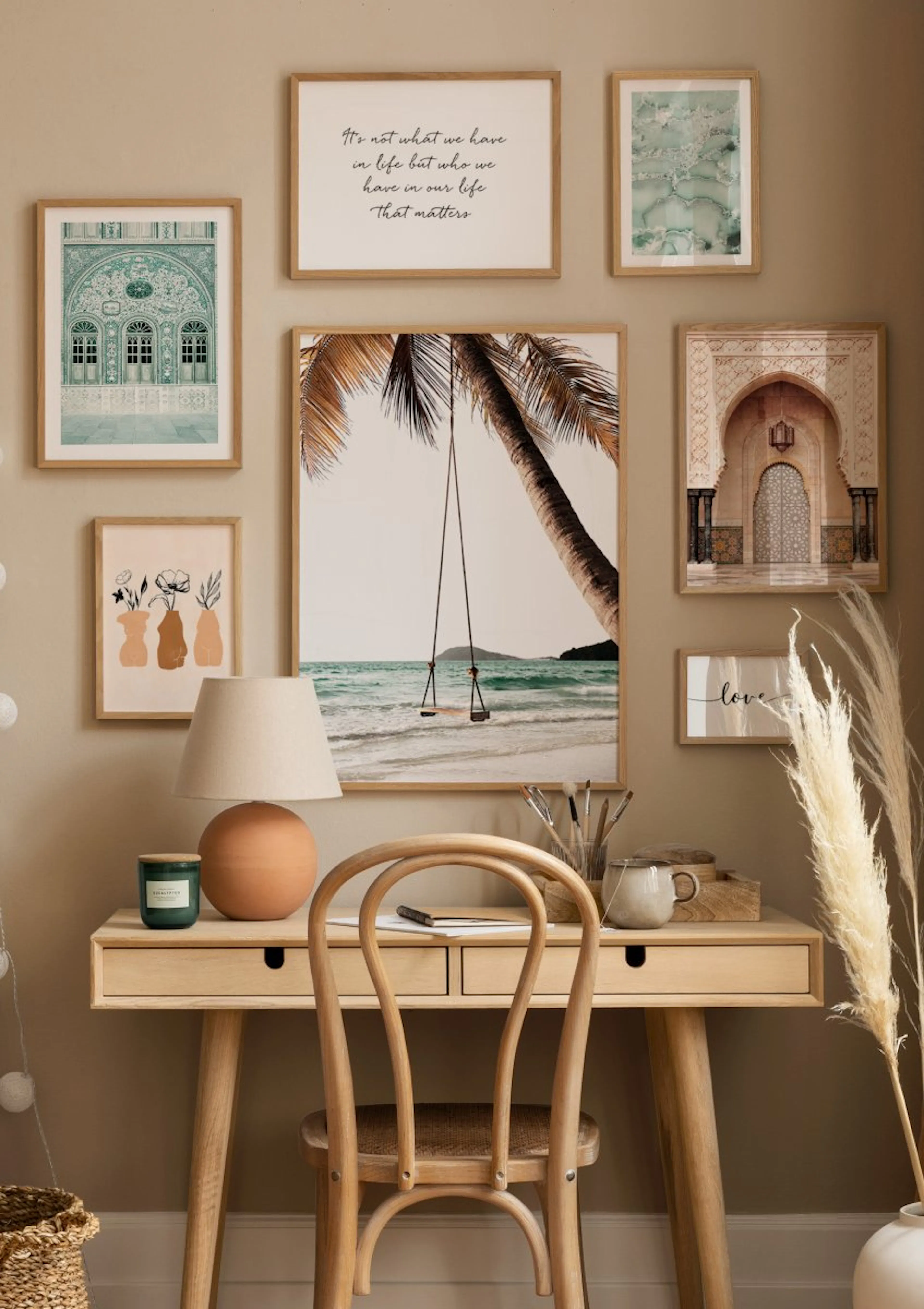A gallery wall of posters and prints above a natural wood desk, featuring a beach scene with a swing, Moroccan architecture, and