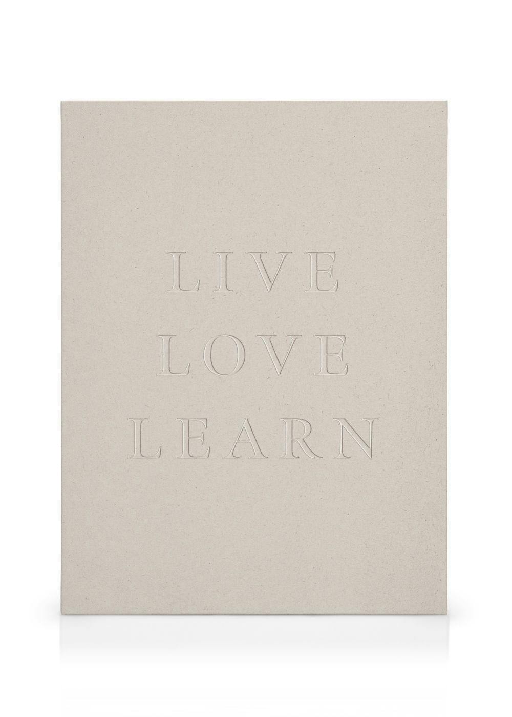 En beige canvastavla med orden LIVE LOVE LEARN på en vit vägg, bredvid en rottingbyrå och trästol.