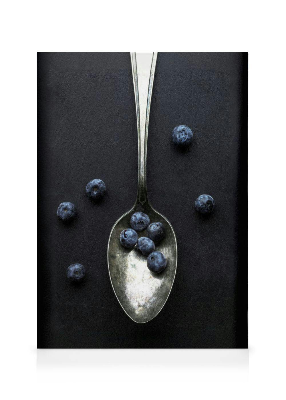 Toile avec une cuillère argentée pleine de myrtilles bleues sur un fond noir texturé, quelques myrtilles dispersées.