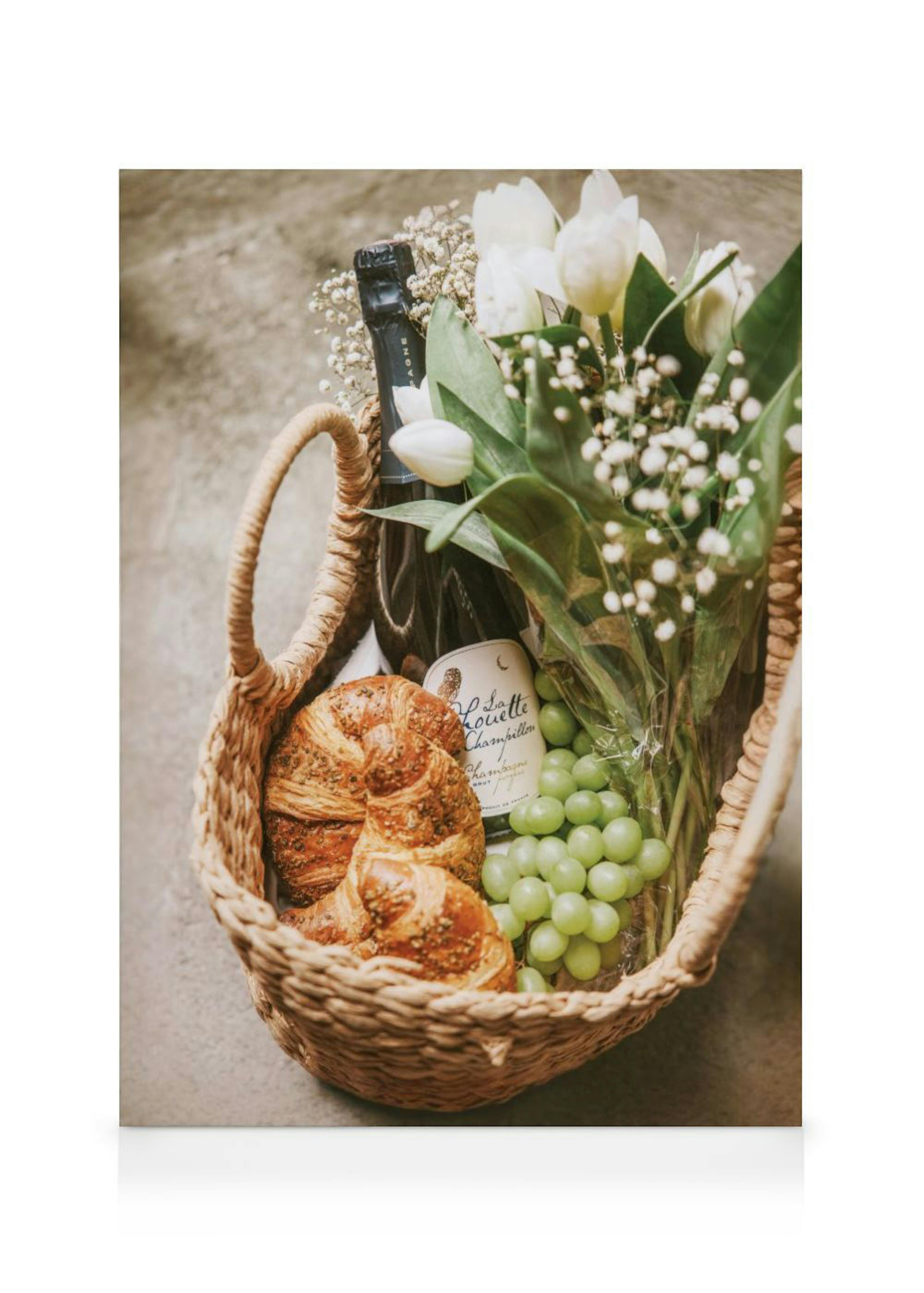 Obraz na płótnie przedstawiający wiklinowy kosz piknikowy z butelką szampana, winogronami, rogalikami i białymi tulipanami.
