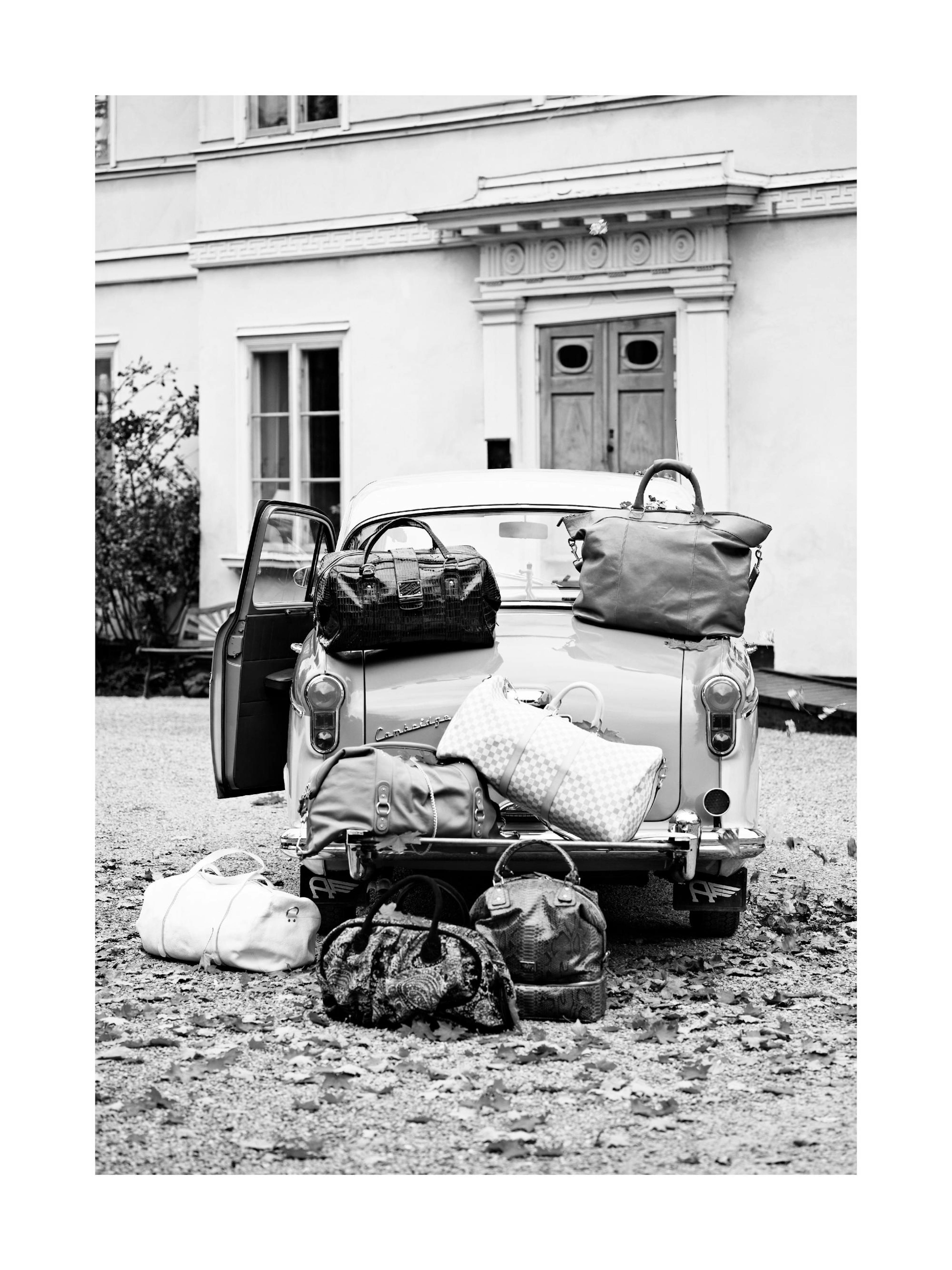 Póster en blanco y negro de un coche clásico con equipaje apilado, frente a una casa.