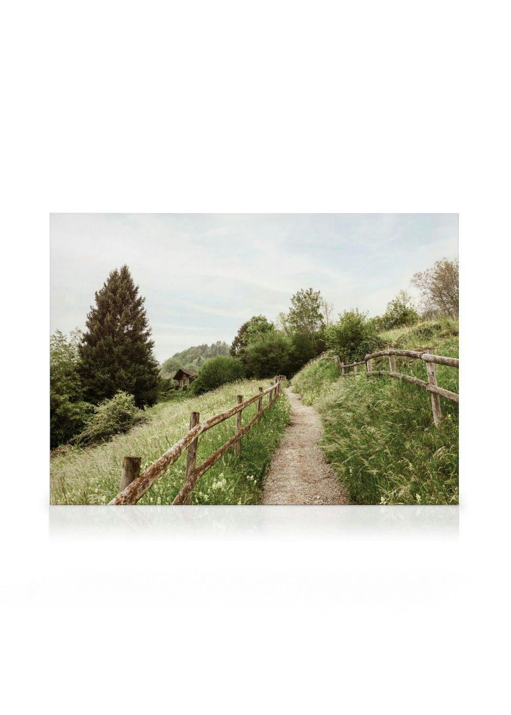 A serene canvas print of a winding path through a grassy field with a rustic wooden fence and distant cabin.
