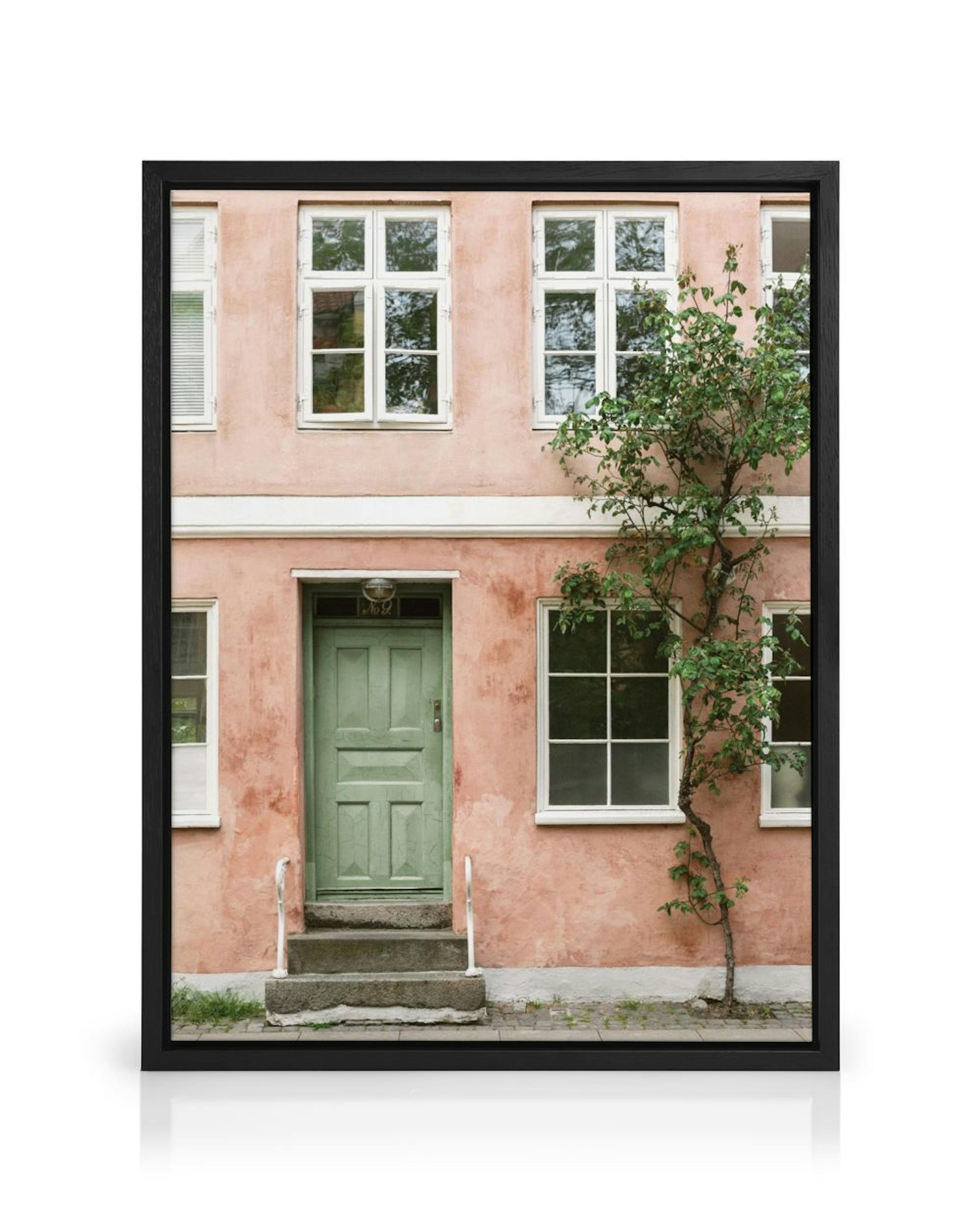 A canvas of a peach building with a green door and white windows, displayed on a white wall above a wooden chair.
