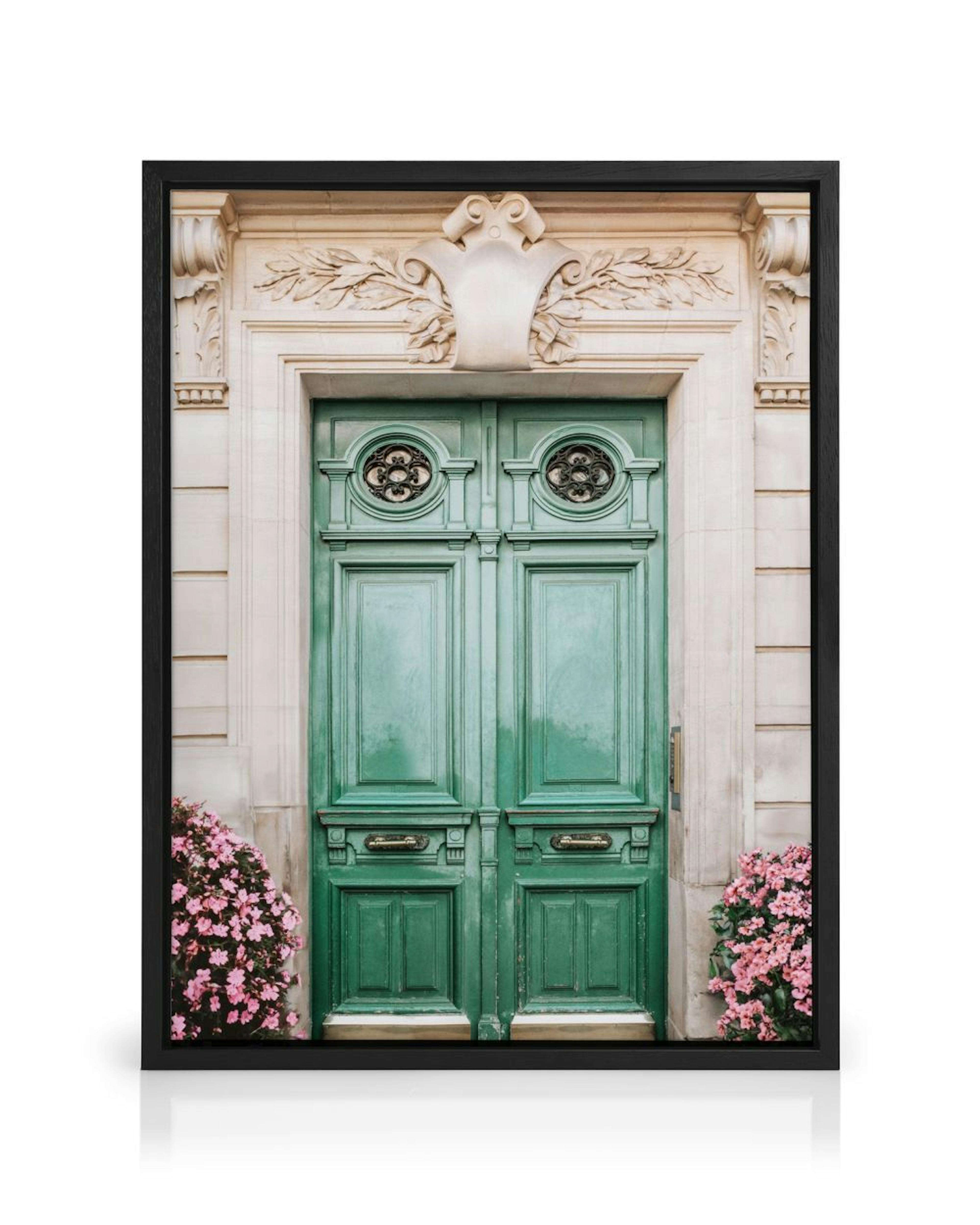 A canvas of a green ornate door with pink flowers, hanging on a light wall above a cane cabinet and wood chair.