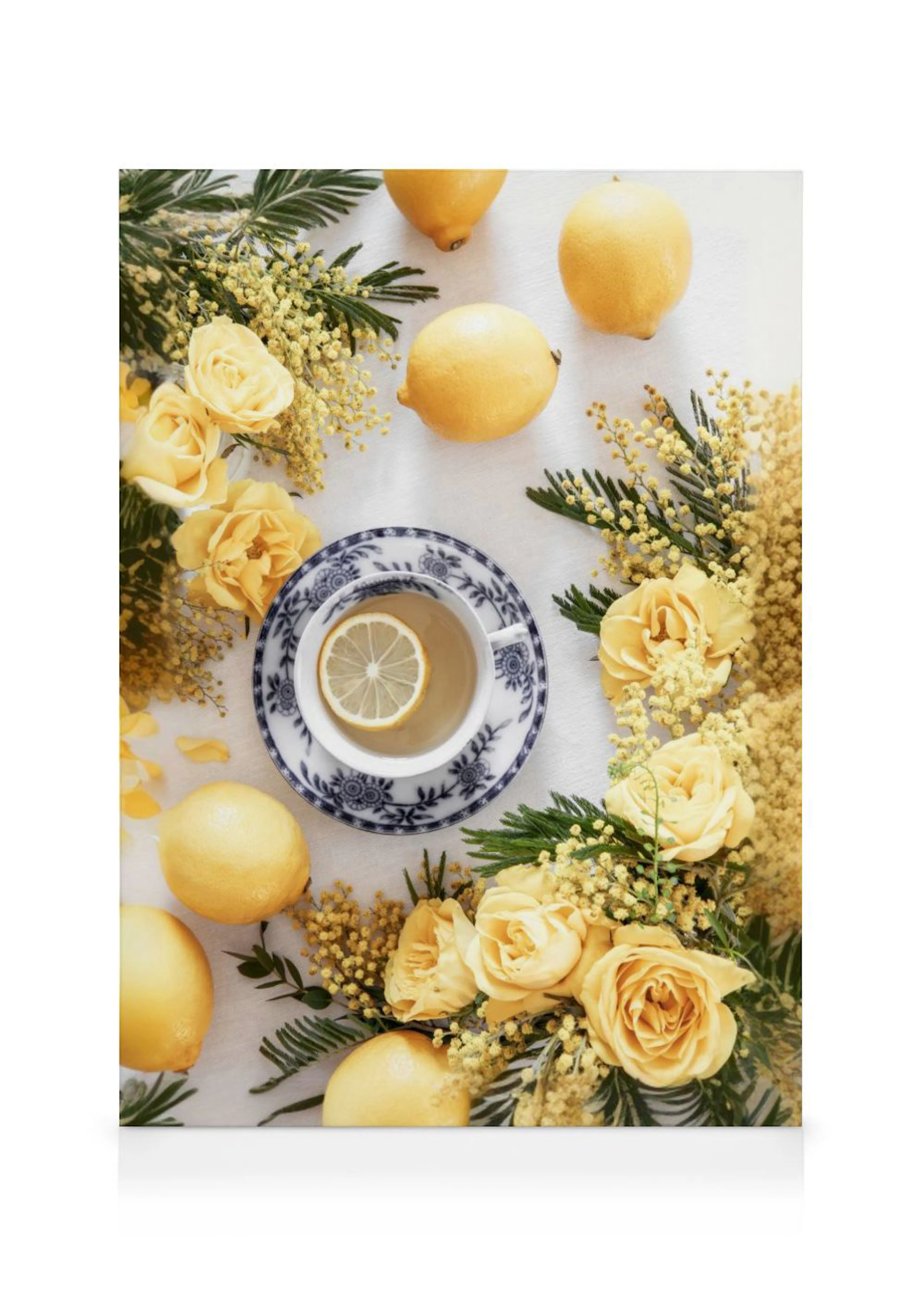En canvastavla med en tekopp, citroner samt guldgula rosor och mimosakvistar på en vit duk.