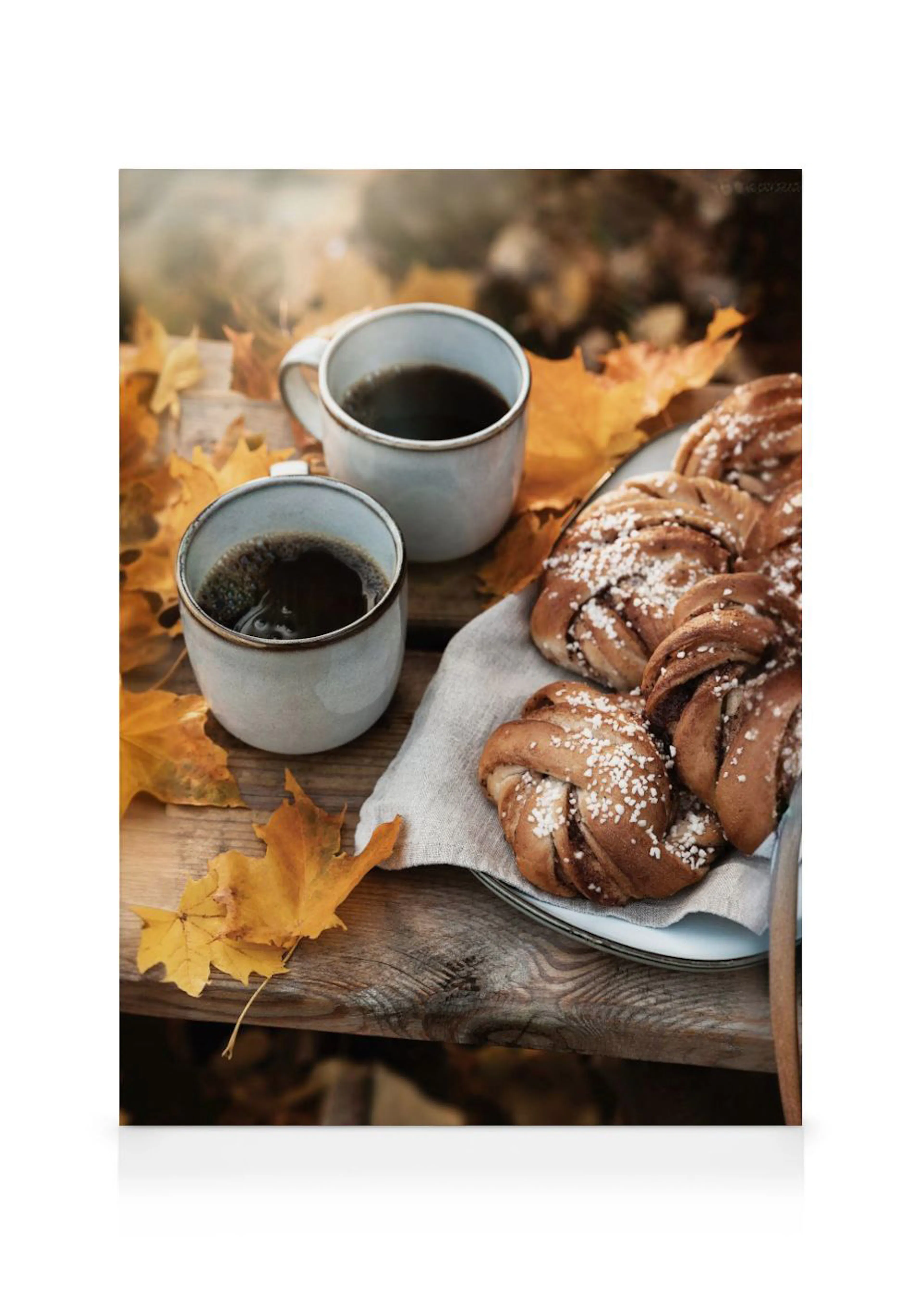 Un lienzo con dos tazas de café y bollos de canela espolvoreados en una mesa de madera con hojas de otoño.