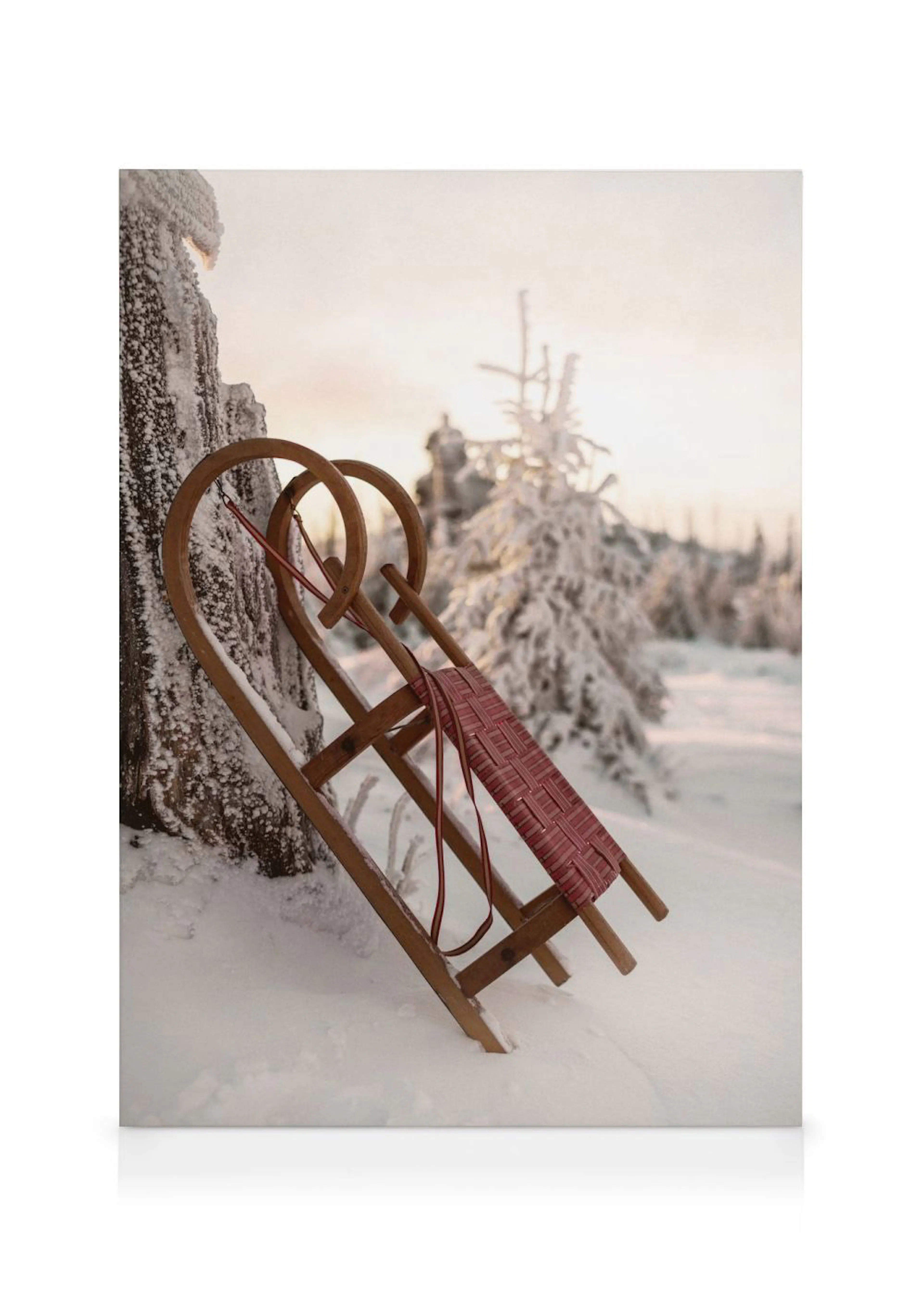 Leinwandbild eines Holzschlittens mit roter Sitzfläche im Schnee, nahe einem schneebedeckten Baum, bei Sonnenuntergang.
