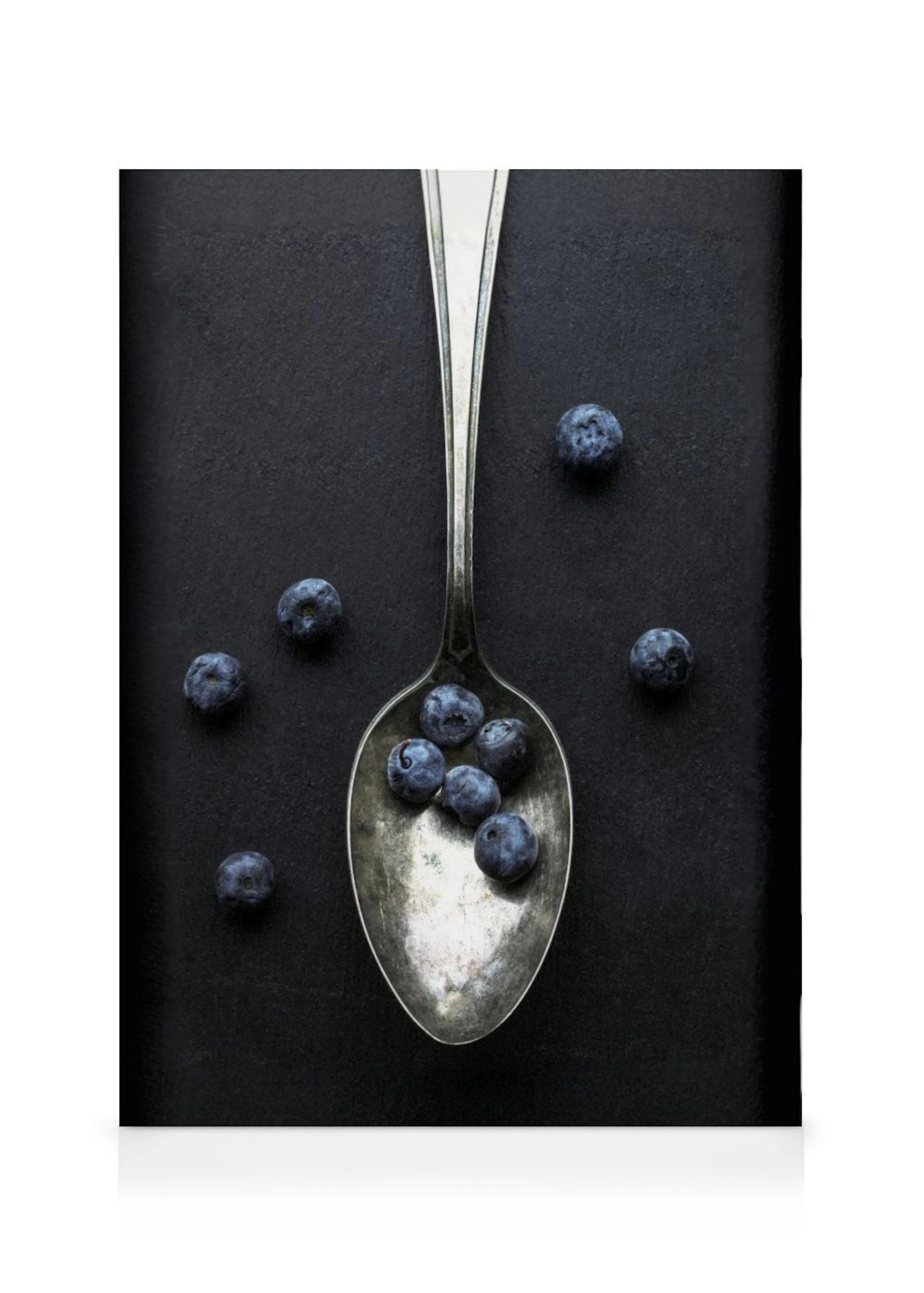 Lienzo con arándanos azules y una cuchara plateada sobre un fondo negro.