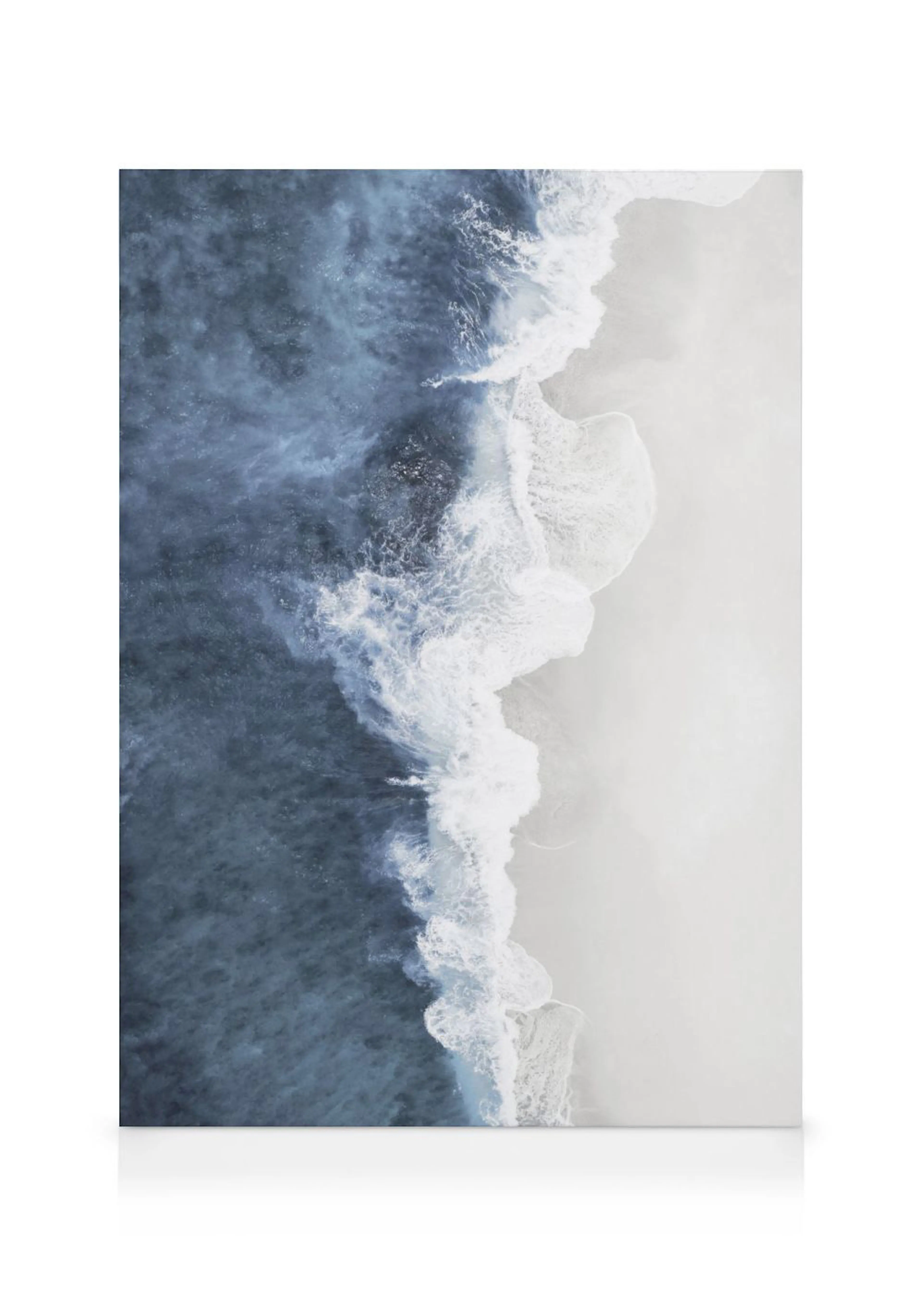 An aerial view canvas of a blue ocean with white foamy waves crashing onto a light sandy beach.