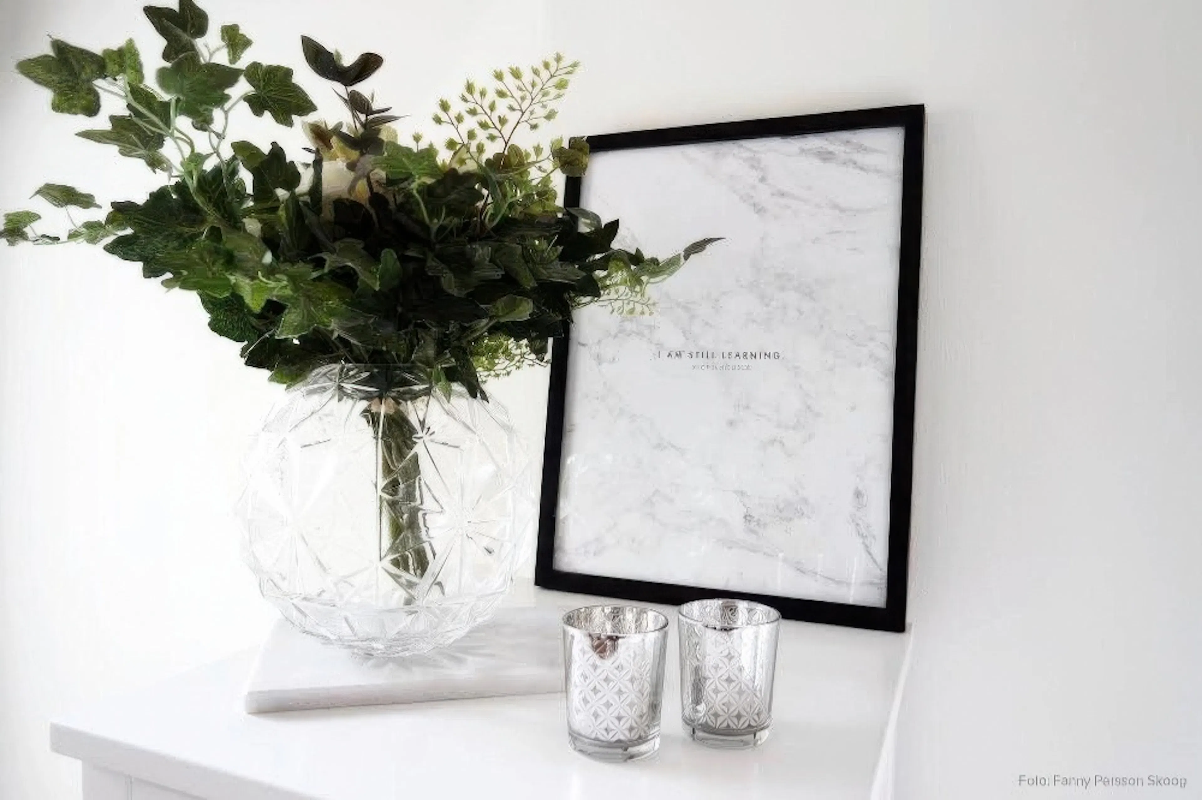 A poster with the text I AM STILL LEARNING on a white marble background, displayed on a white shelf with a glass vase and two ca