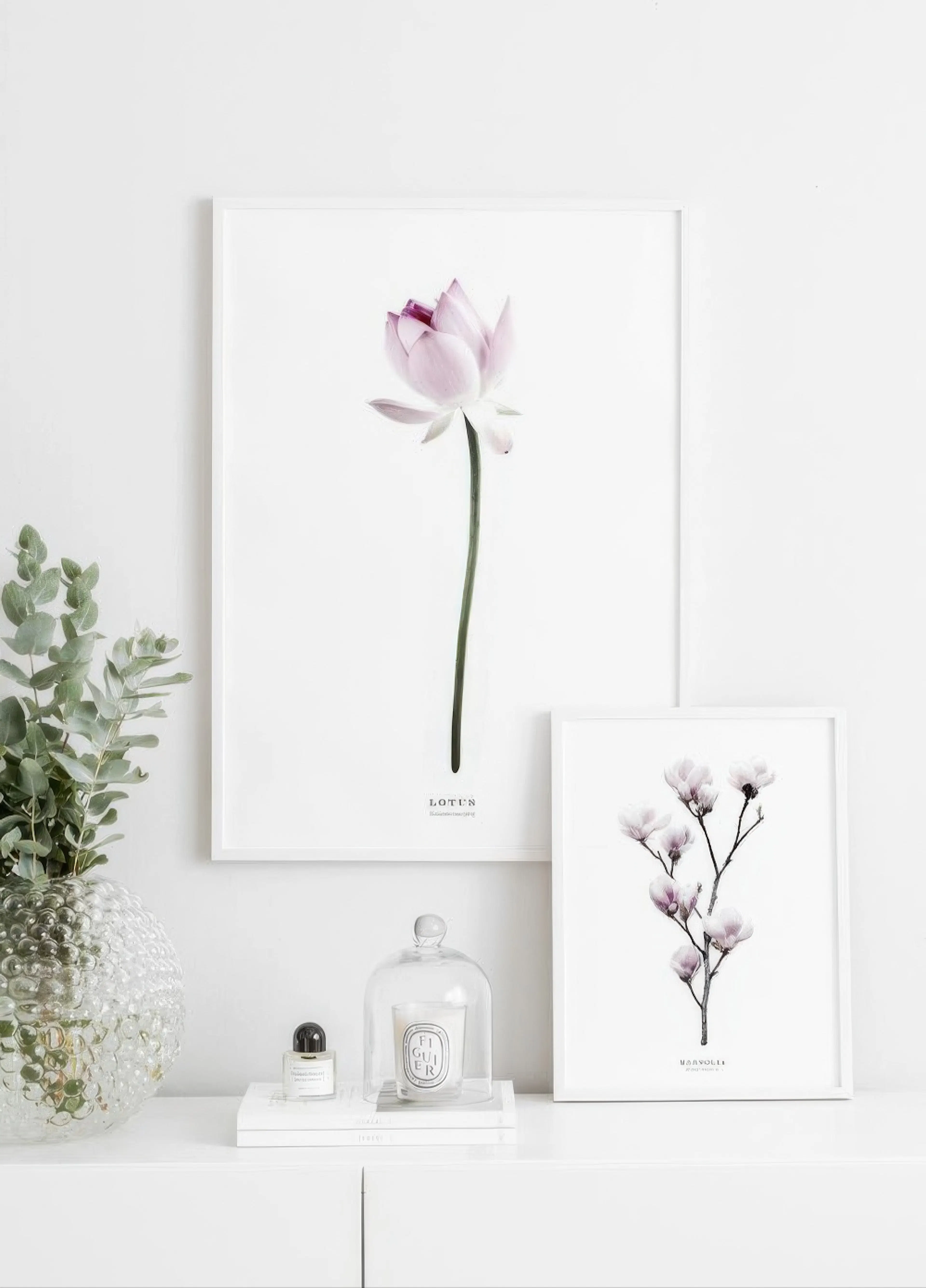 Two botanical posters of a pink lotus and light purple magnolia branch displayed on a white console table with a plant and candl