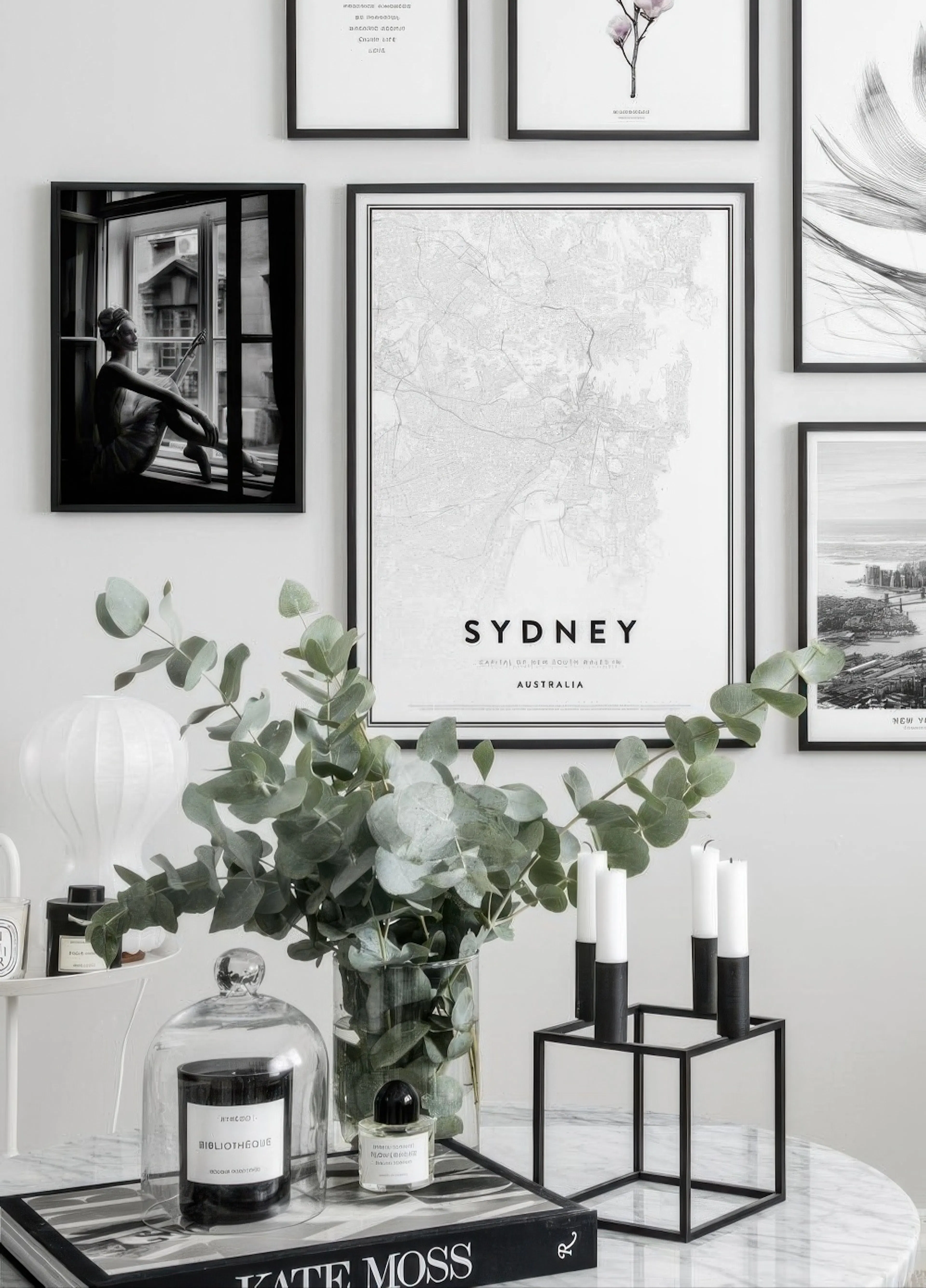 A poster of a black and white Sydney map, displayed in a gallery wall above a marble table with eucalyptus, candles, and books.