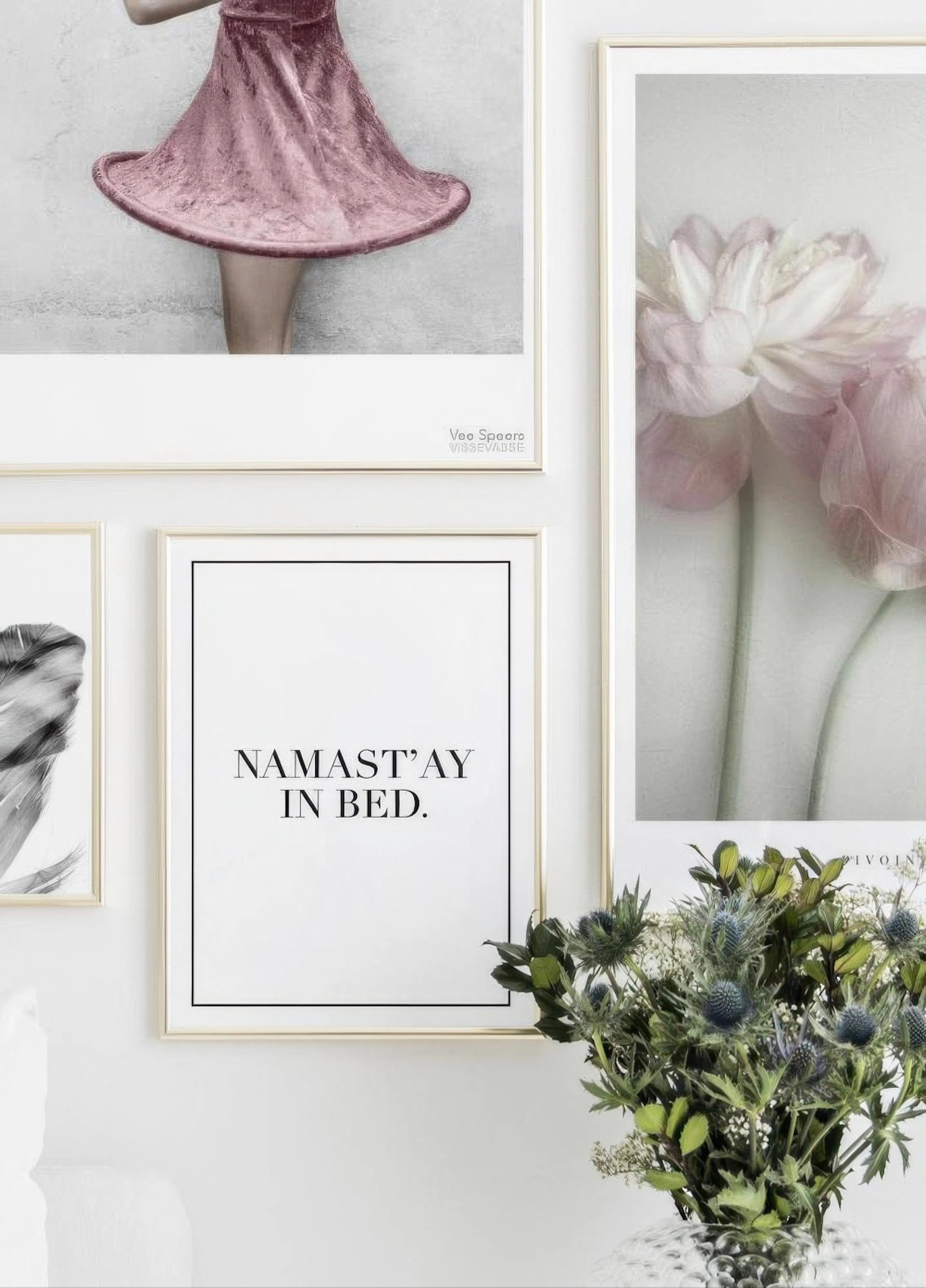 A gallery wall with three posters, including a pink dress, pink flowers, and Namastay In Bed text, with a vase of thistles.