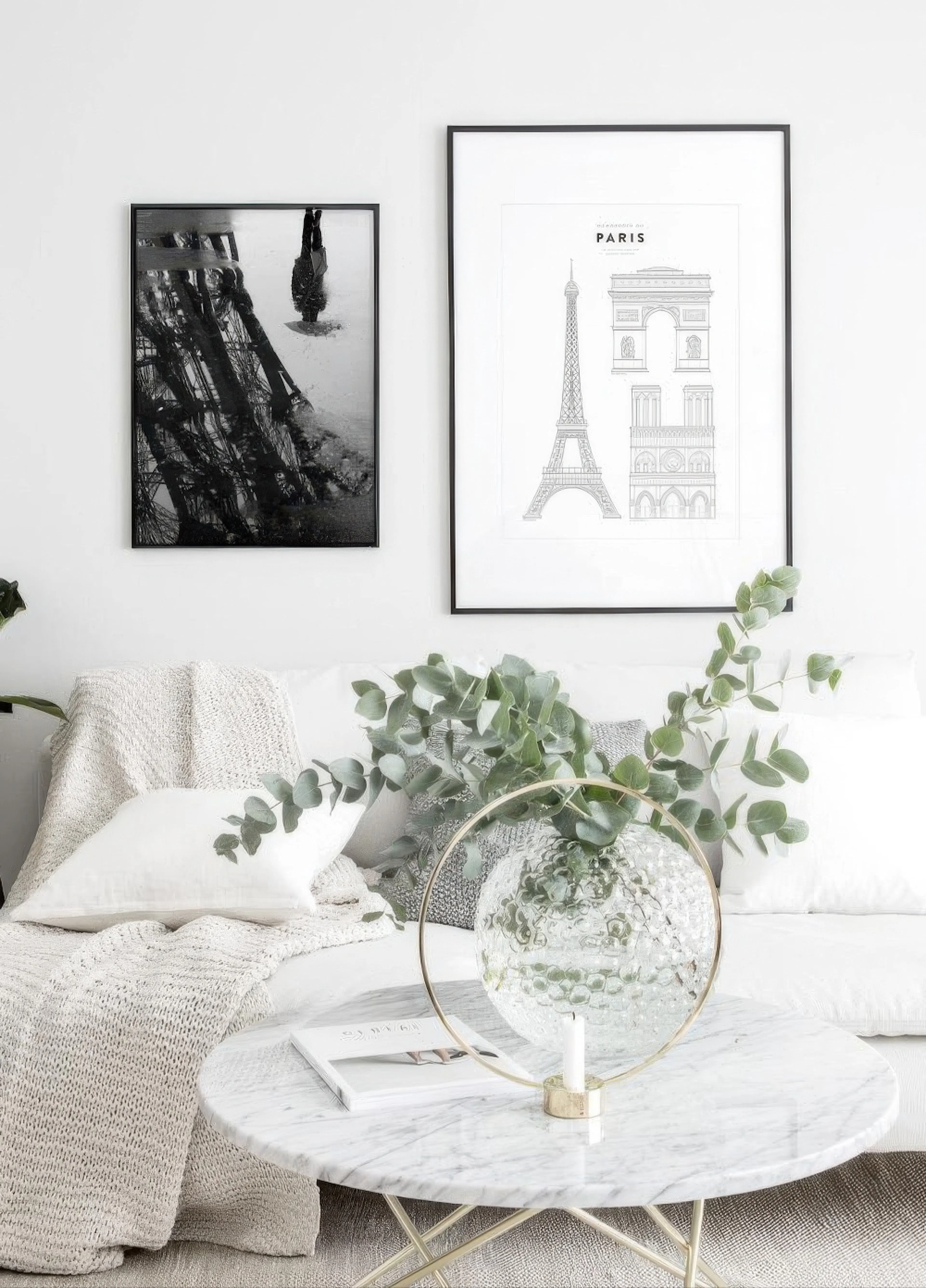 Kaksi julistetta roikkuu valkoisella seinällä sohvan yläpuolella. Vasemmalla mustavalkoinen Eiffel-tornin heijastus, oikealla Pa