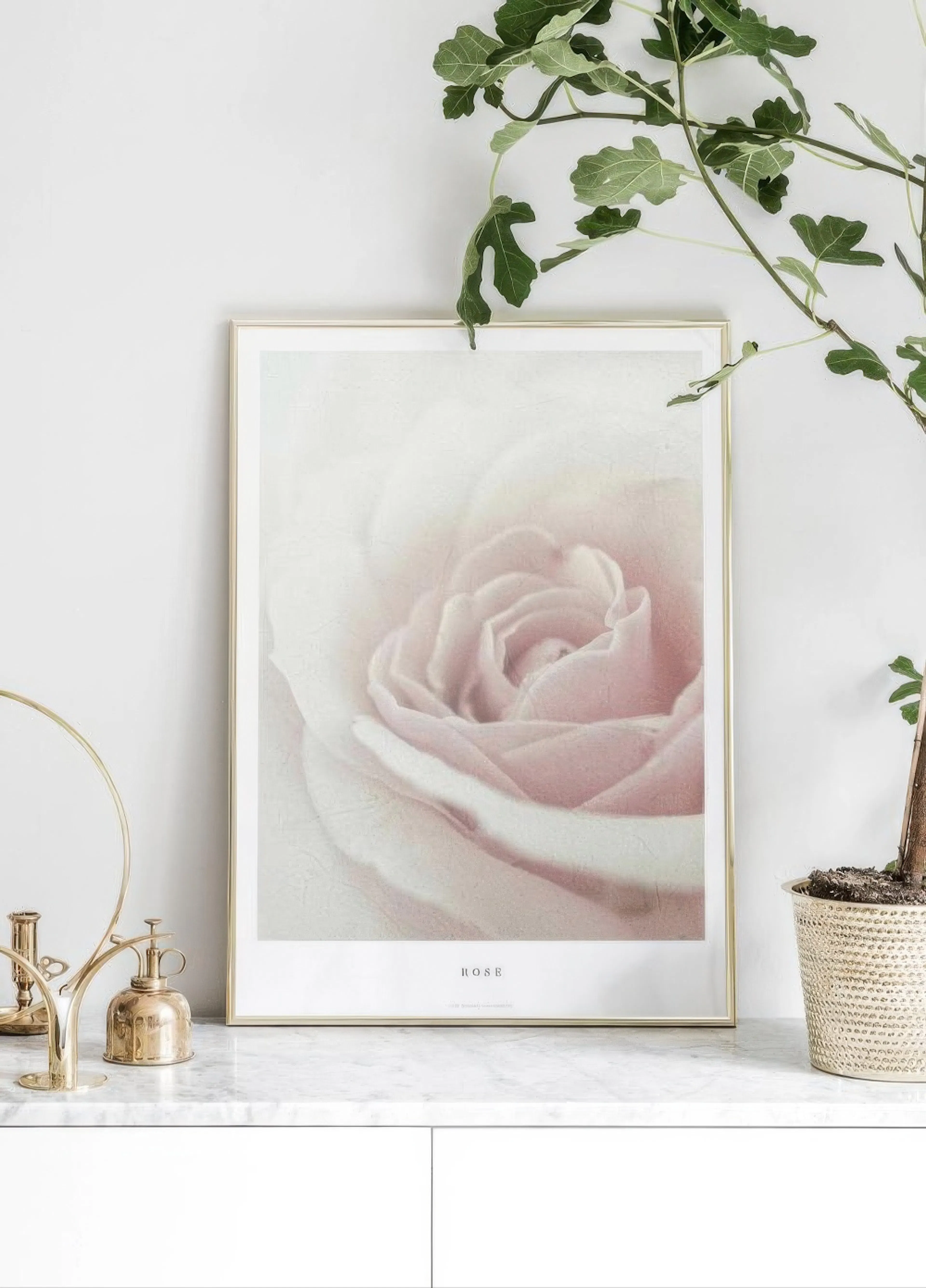 A poster featuring a delicate pink rose close-up, displayed on a marble surface with a plant and brass decor.