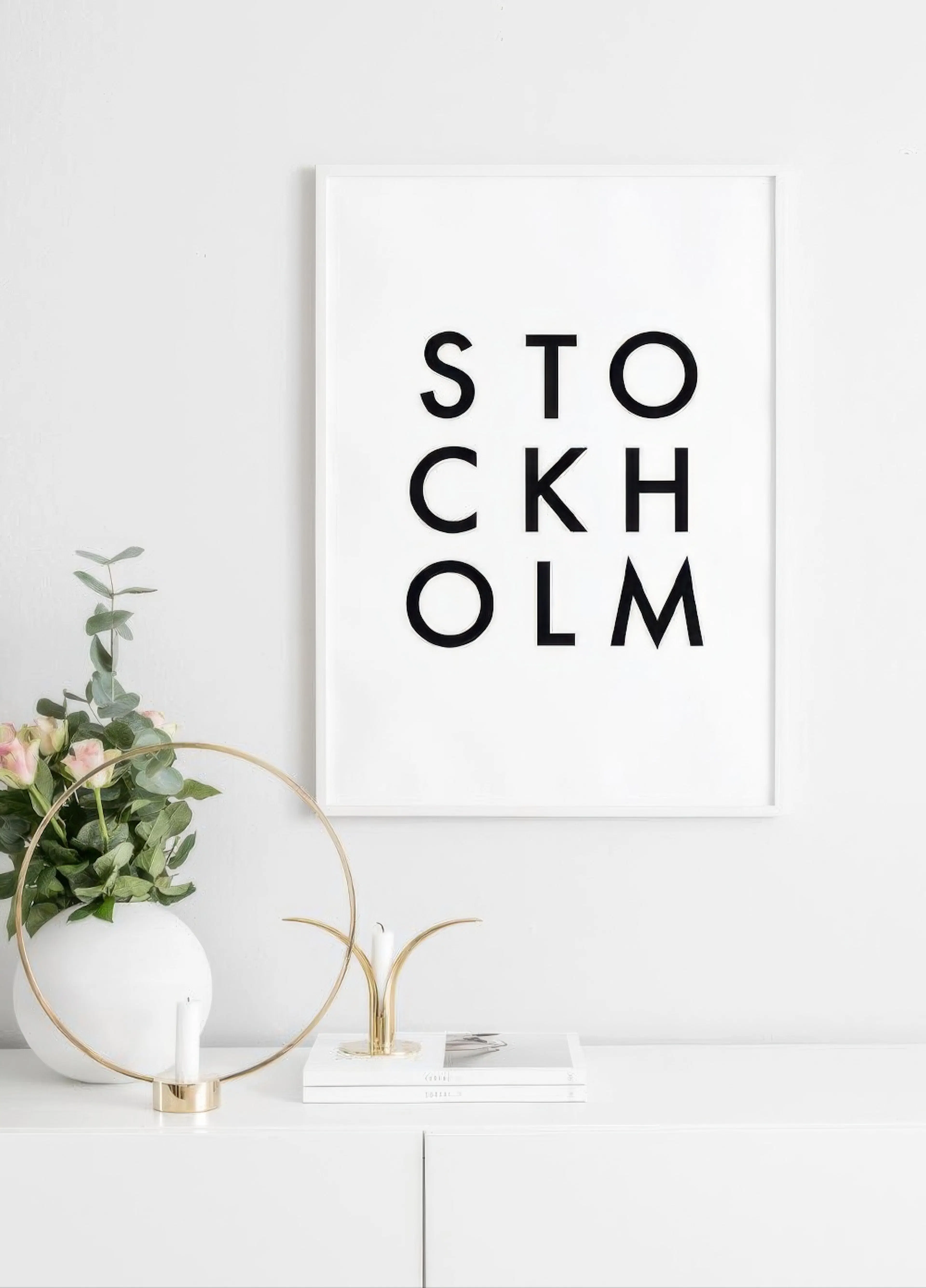 A black and white typography poster spelling Stockholm, displayed above a white cabinet with a vase of flowers and candles.