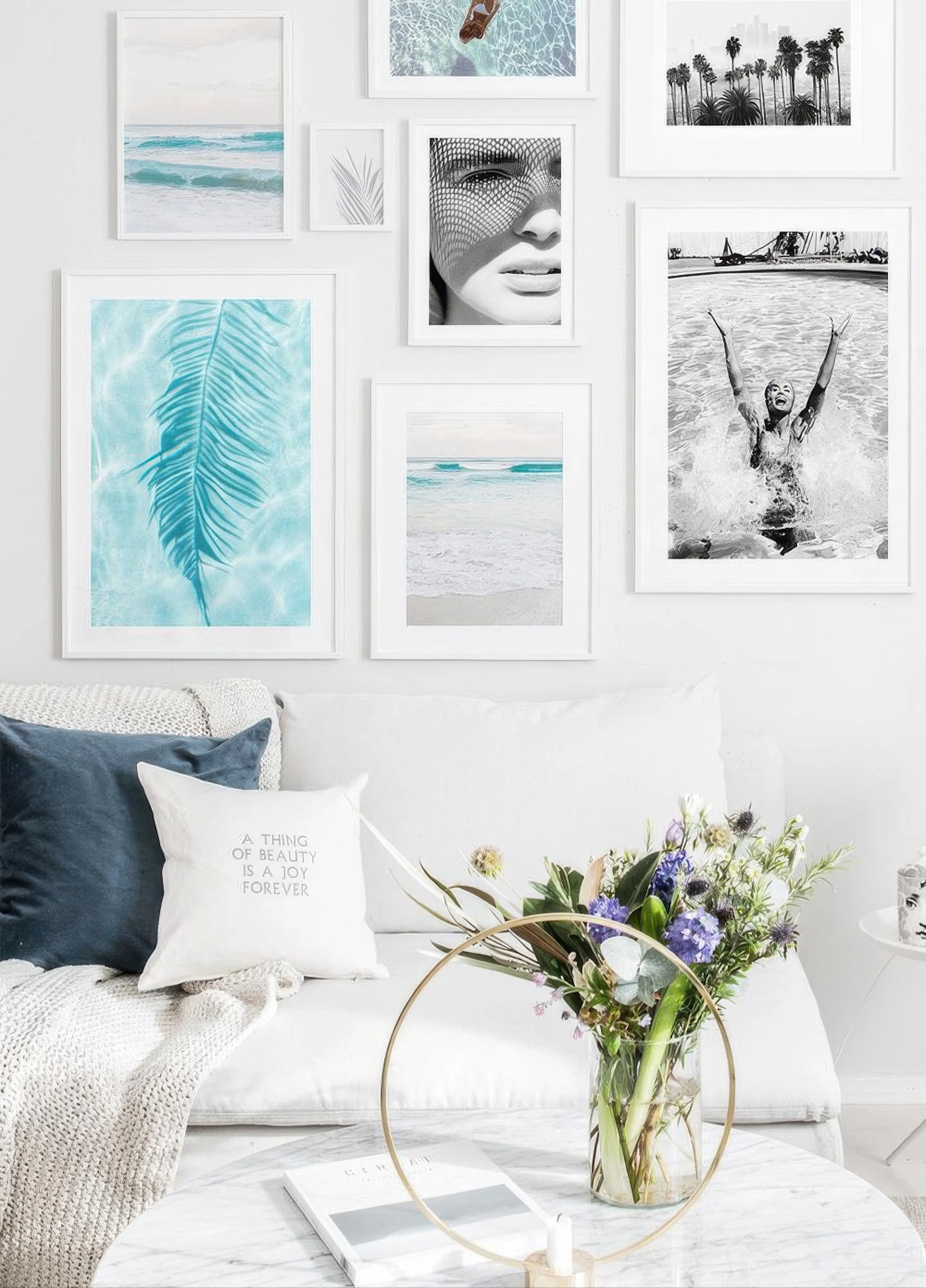 Julistetauluseinä valkoisella seinällä, jossa on merimaisemia, palmunlehtiä ja mustavalkokuvia. Alla valkoinen sohva ja marmorip