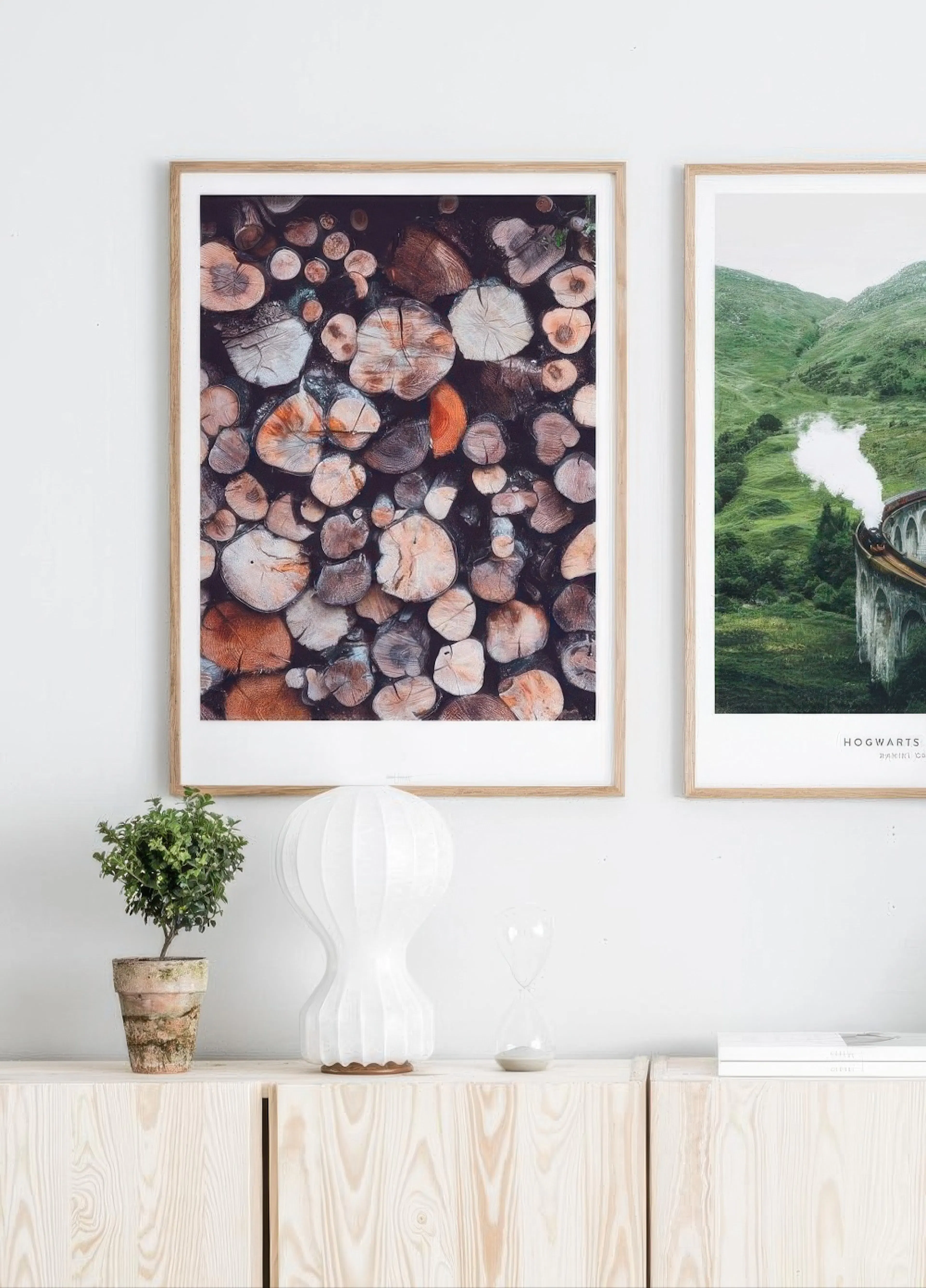 Plakát s detailním pohledem na hromadu nařezaného dříví, zavěšený nad dřevěnou komodou s rostlinou a lampou.