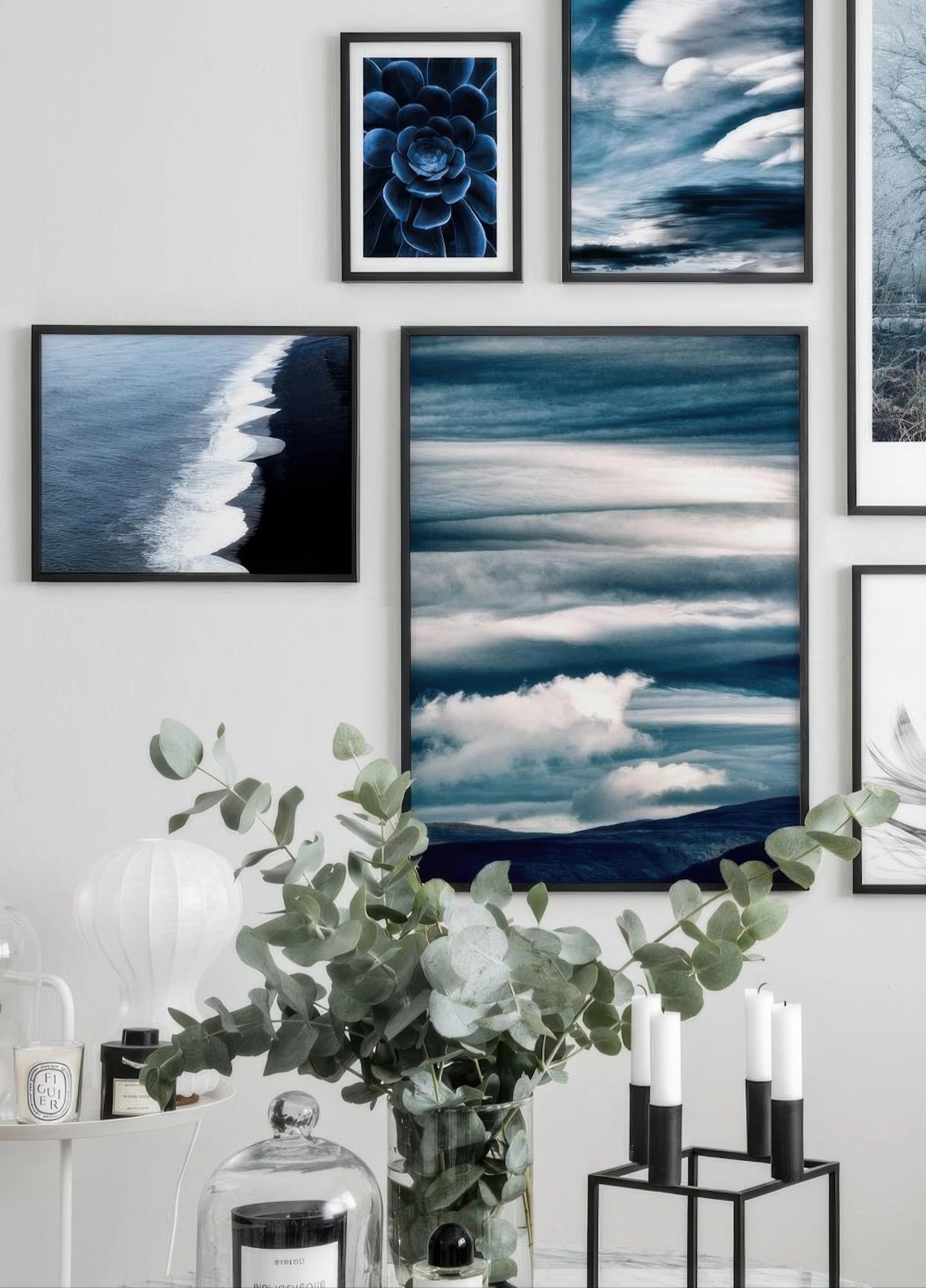 Nástěnná galerie modrých plakátů s přírodními motivy: sukulent, moře a obloha, nad eukalyptem ve váze a svíčkami.