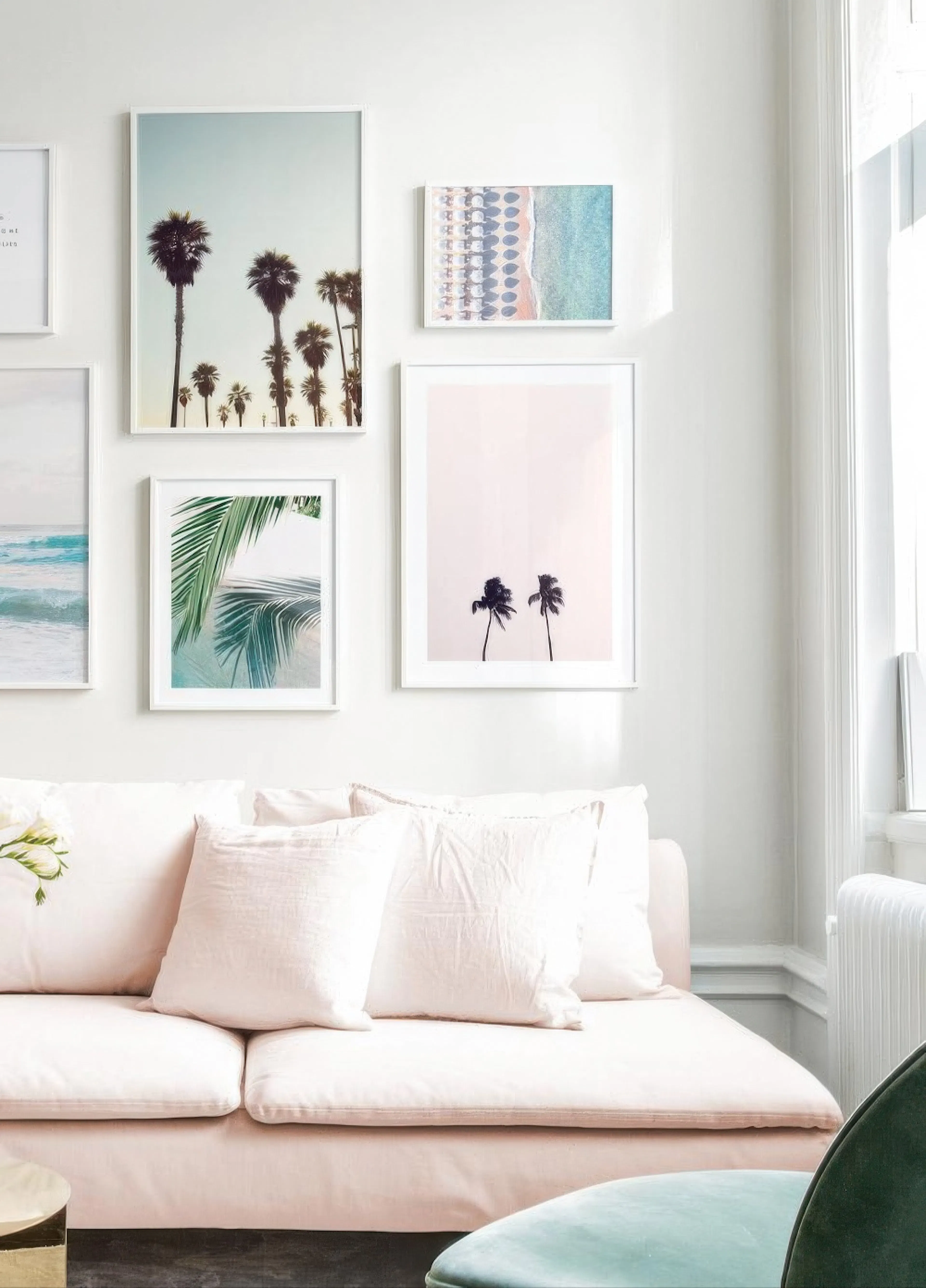 Nástěnná galerie plakátů s palmami a pláží visící nad růžovou pohovkou s bílými polštáři v obývacím pokoji.