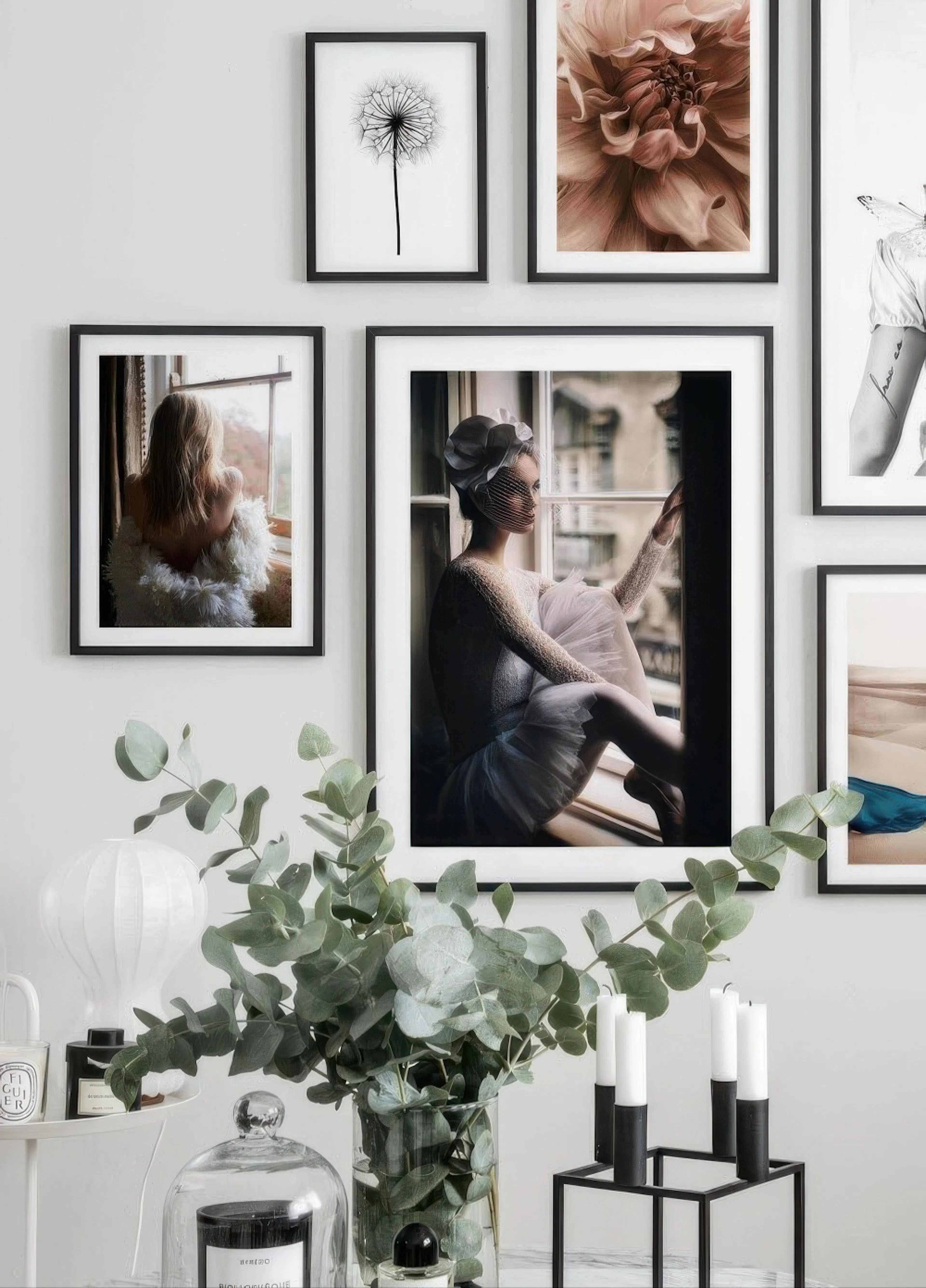 Plakát baletky sedící na parapetu, na stěně s dalšími obrazy. V popředí eukalypt a svíčky.