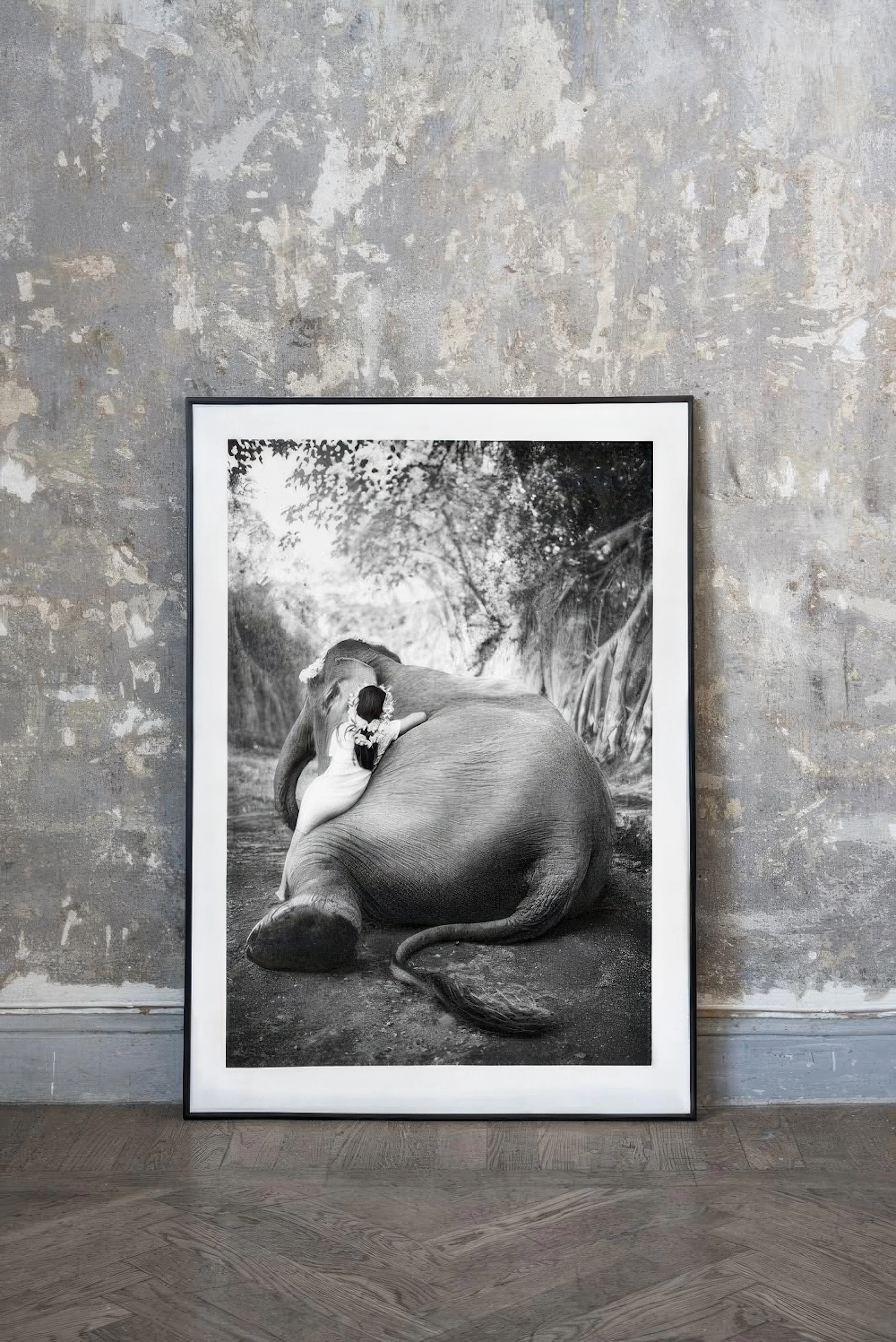 A black and white poster of a person hugging an elephant, displayed on a wooden floor against a textured concrete wall.