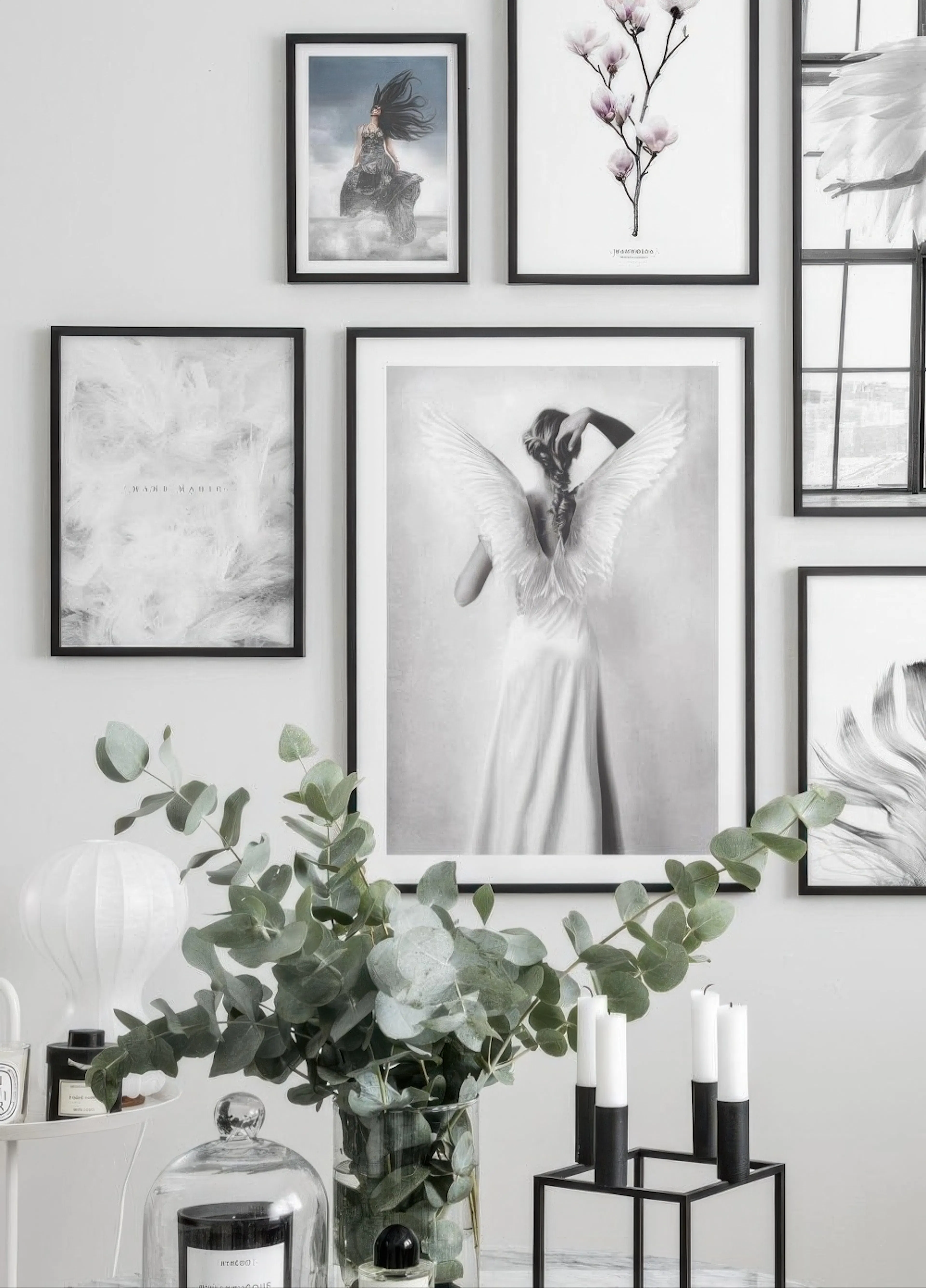 A gallery wall featuring a black and white poster of a woman with angel wings, alongside other prints, above a glass vase with e
