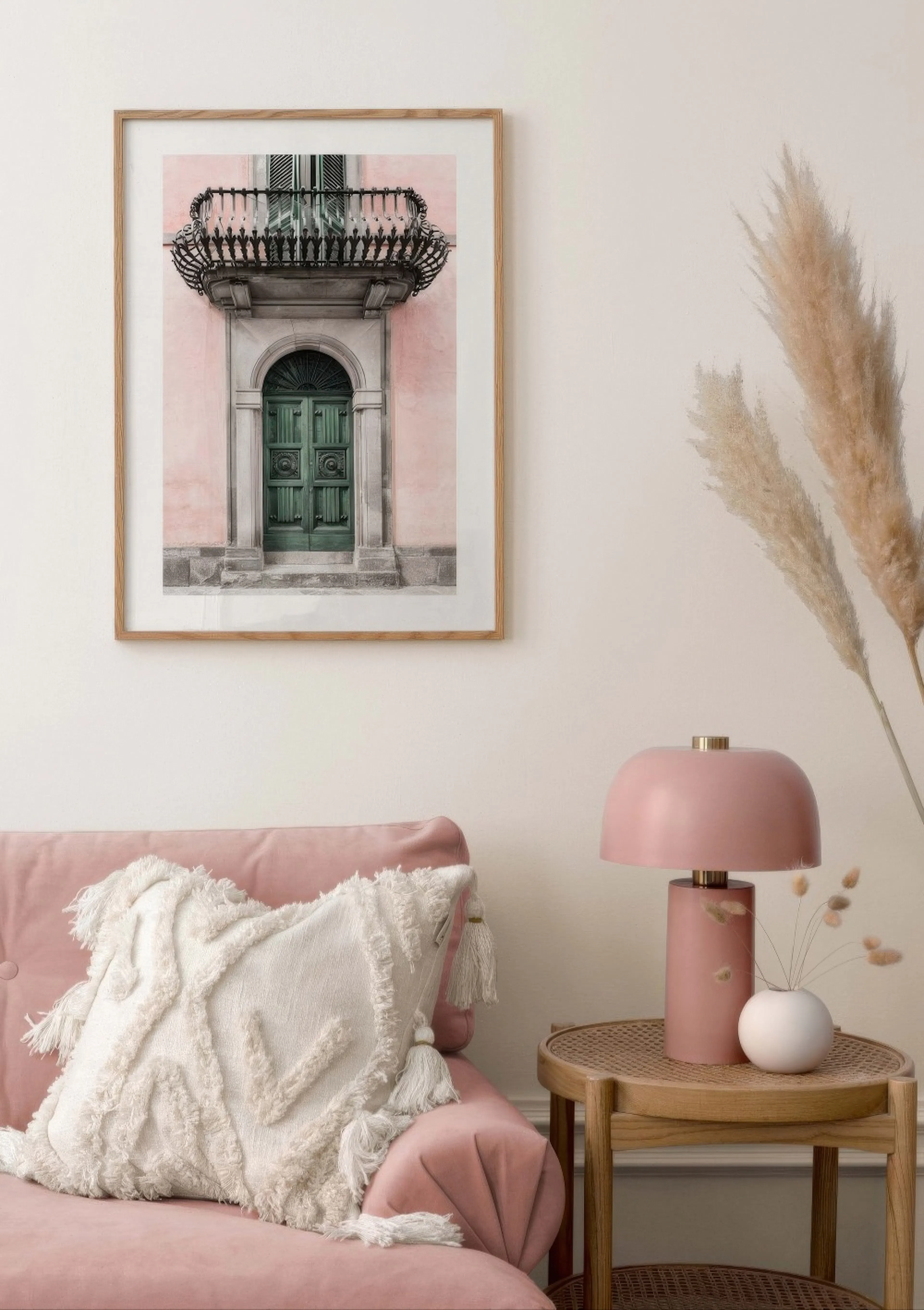 A poster of an arched green door and balcony on a pink building, hanging above a pink sofa with a white cushion.