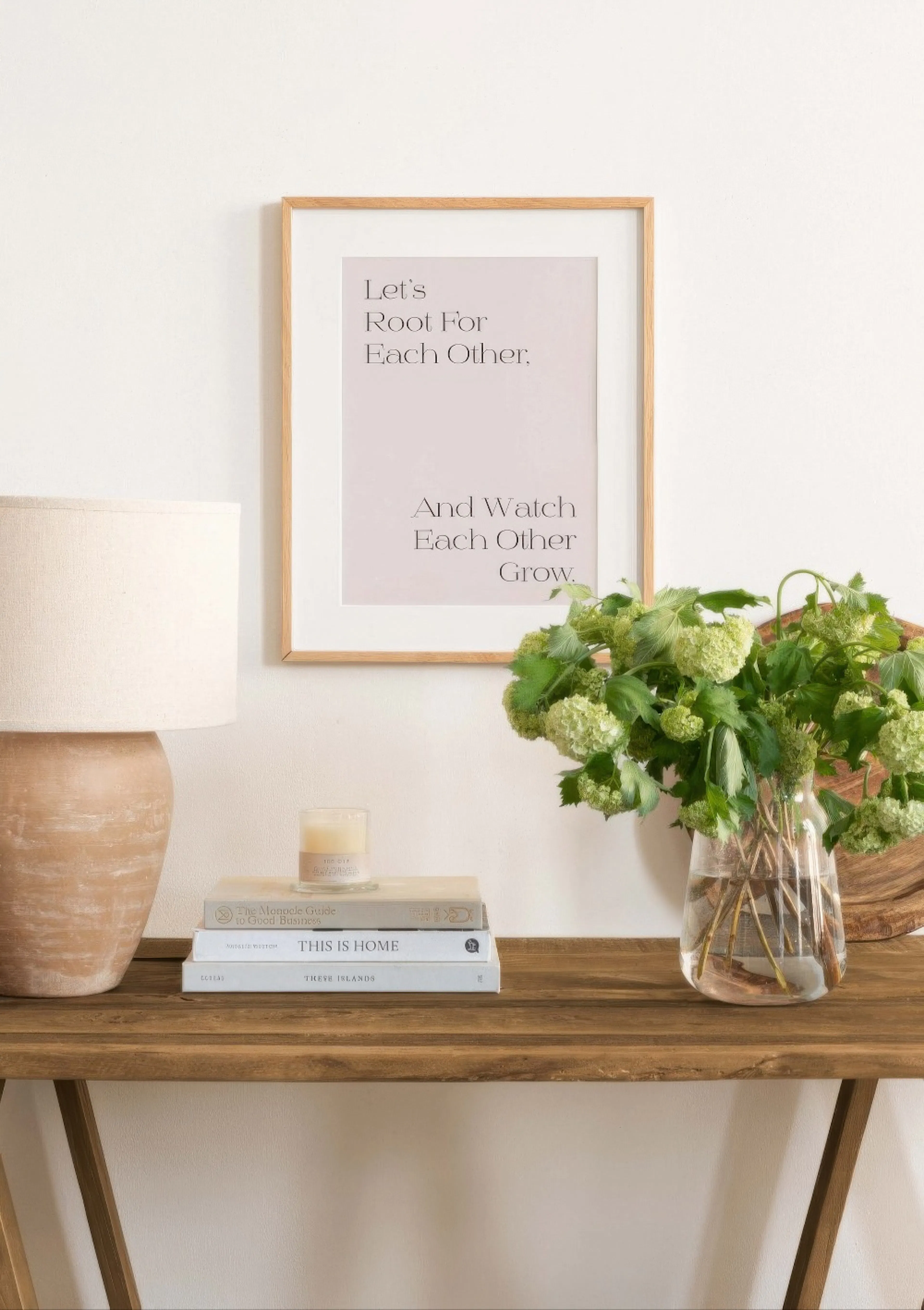 A framed poster with the text Lets Root For Each Other. And Watch Each Other Grow. displayed above a wooden console table with a