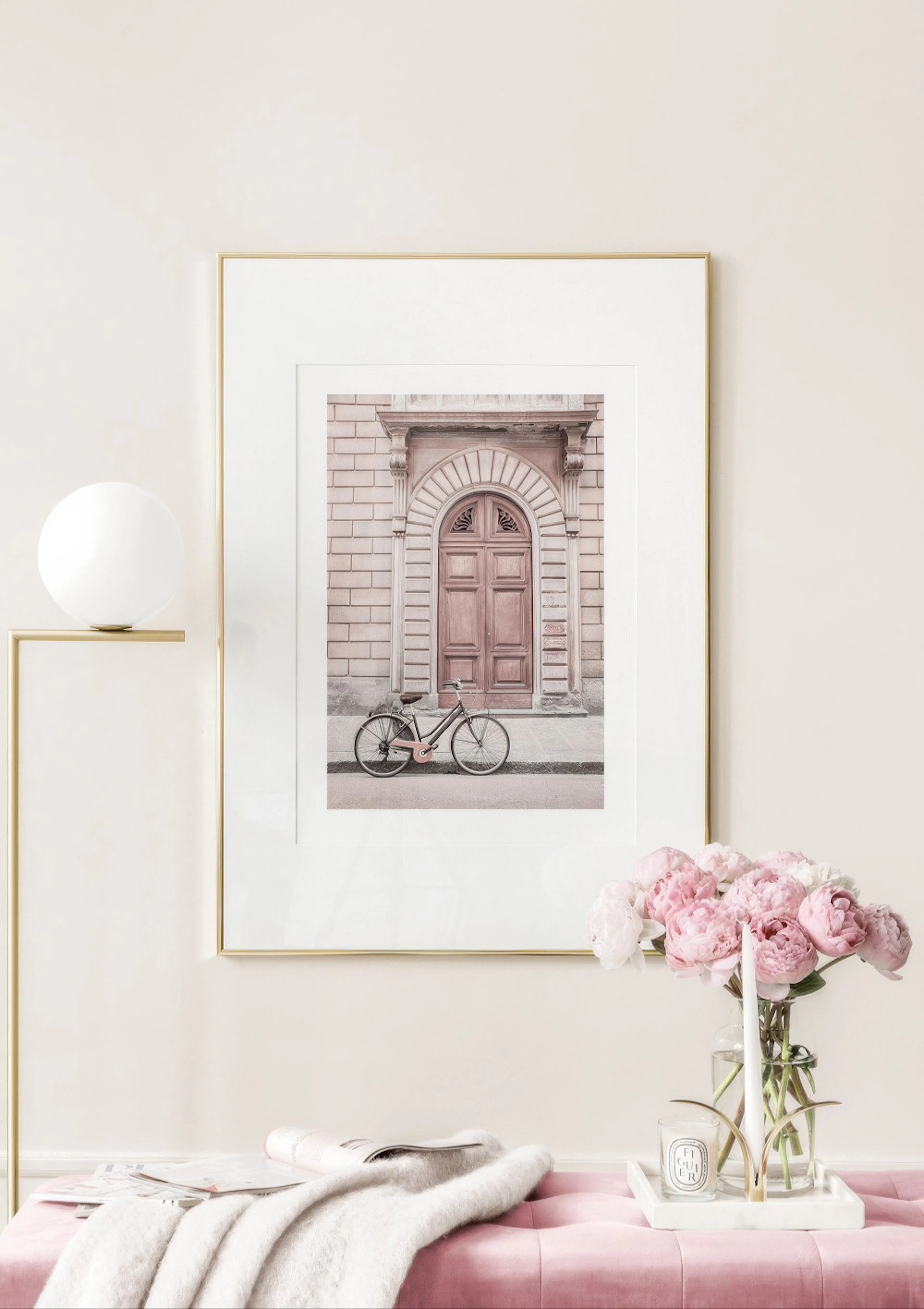 薄ピンクの壁に飾られた、茶色の扉と自転車が写るポスター。手前にはピンクの布張りのベンチ、花瓶に入ったピンクの芍薬、白いキャンドルや本が置かれている。