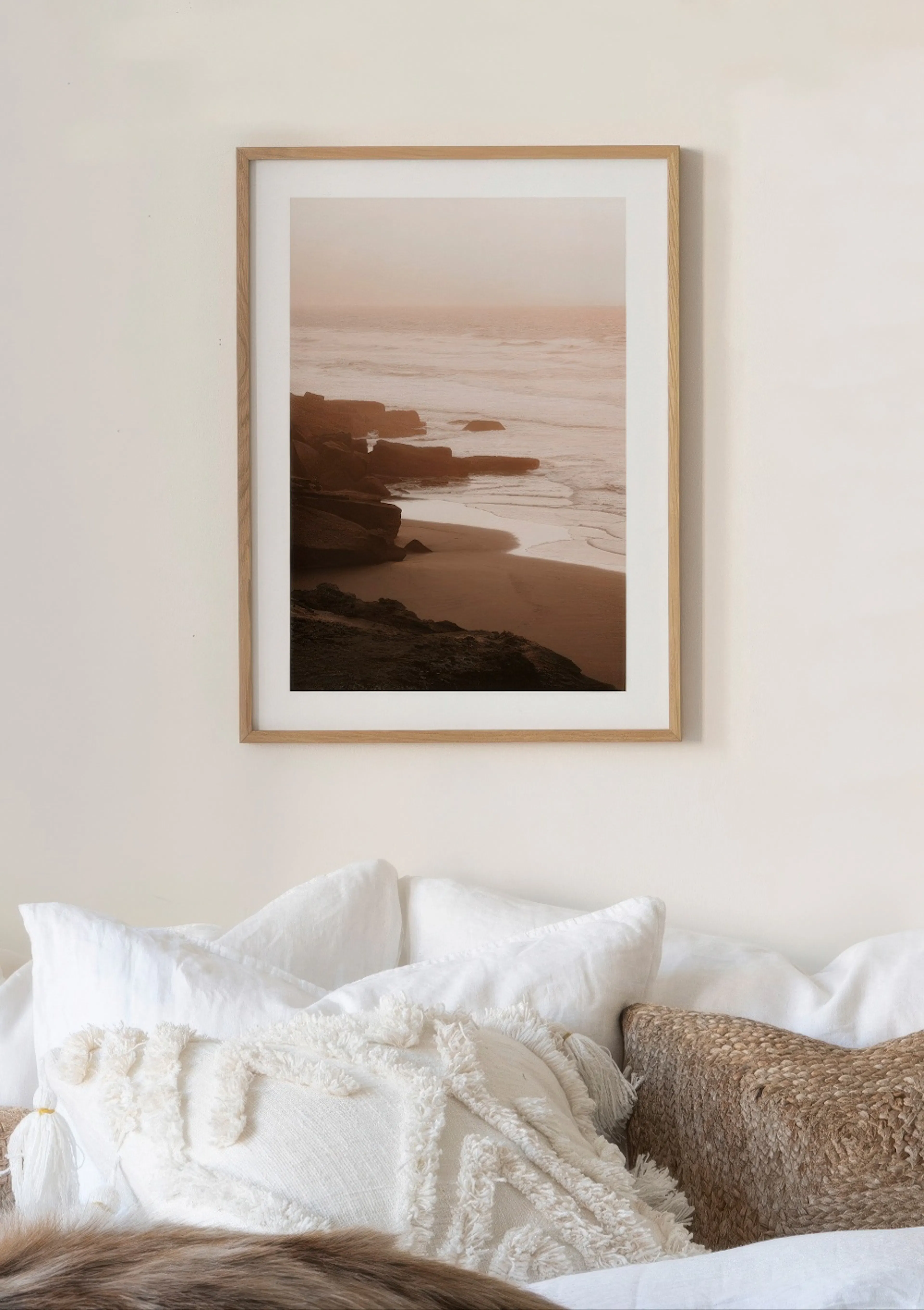 Poster van een zonsondergang aan de kust met rotsen en golven, hangend boven een bed met witte kussens en een decoratief kussen.