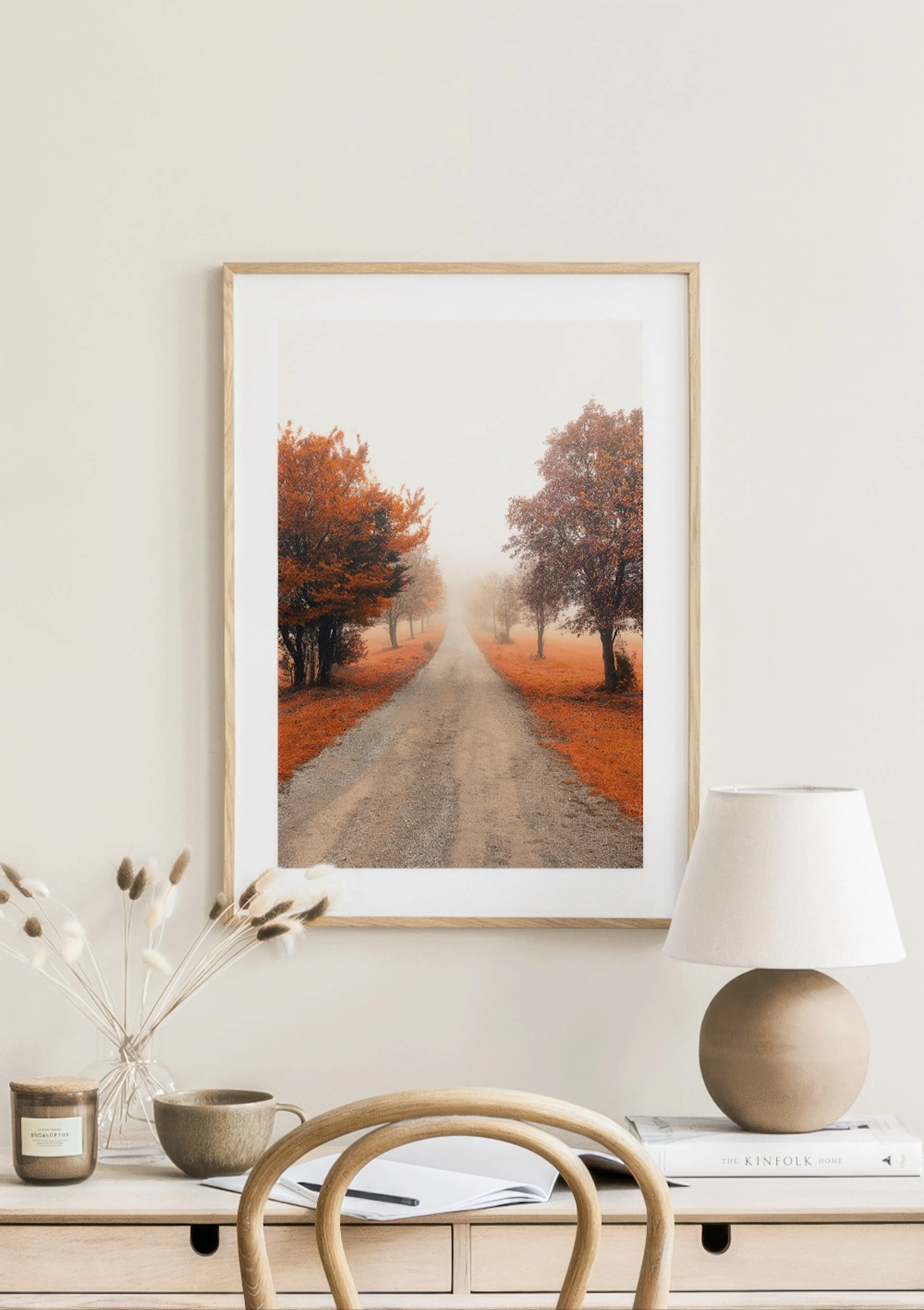 A poster of a hazy country road lined with trees in vibrant autumn colours, displayed above a light wooden desk with a lamp and 